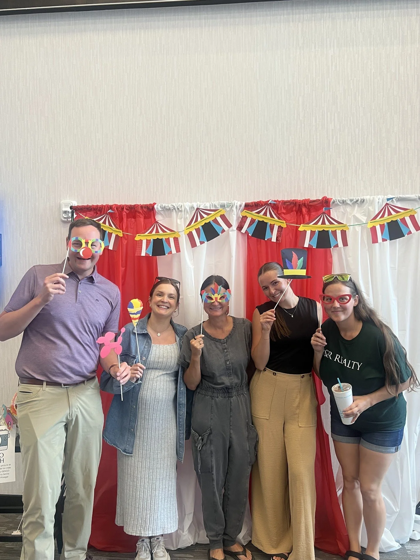 Five people standing in front of a red and white circus-themed backdrop, wearing fun props and holding photo booth accessories at a celebration or party.