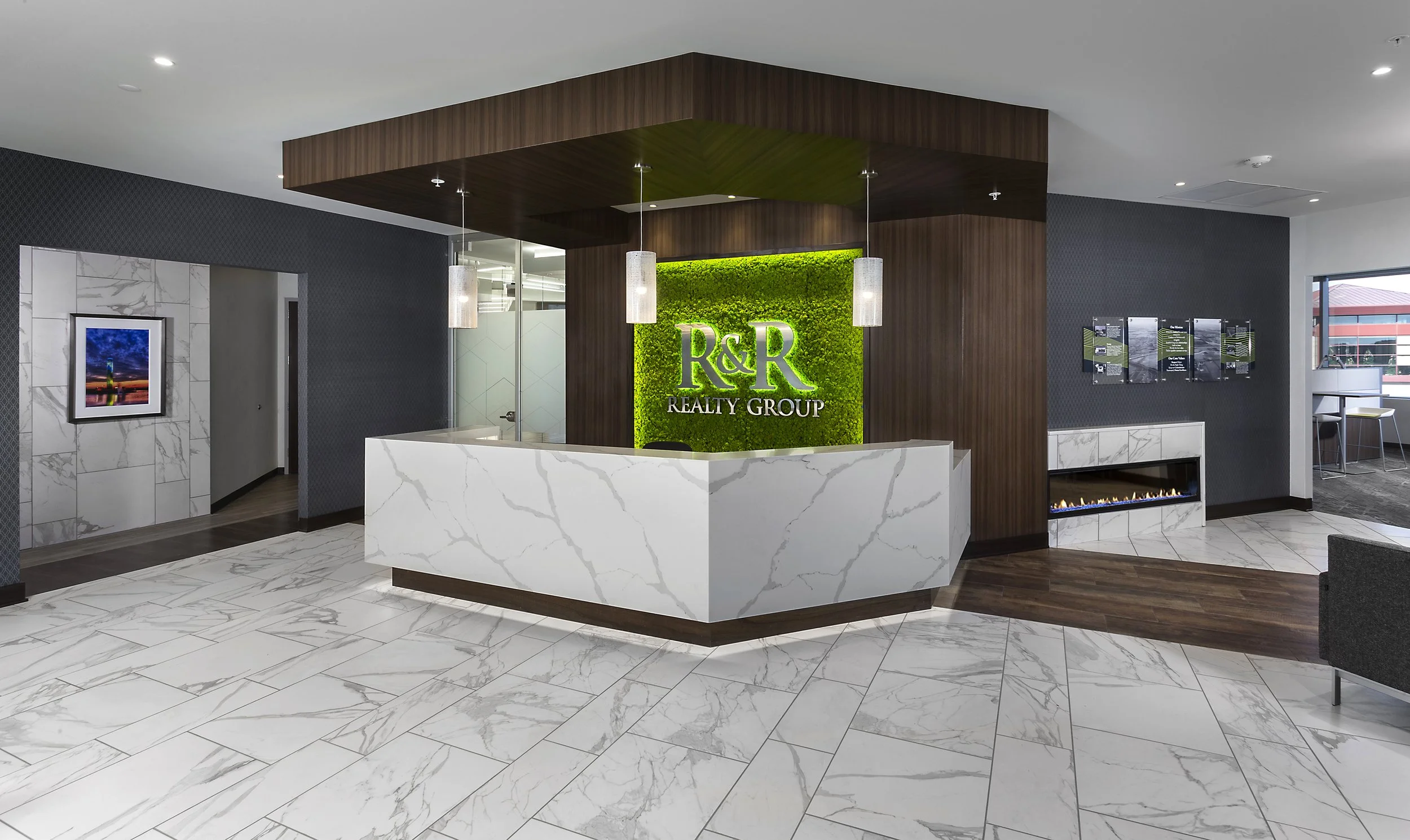 Modern real estate office reception area with R&R Realty Group logo on a green moss wall, white marble desk, and wood accents.