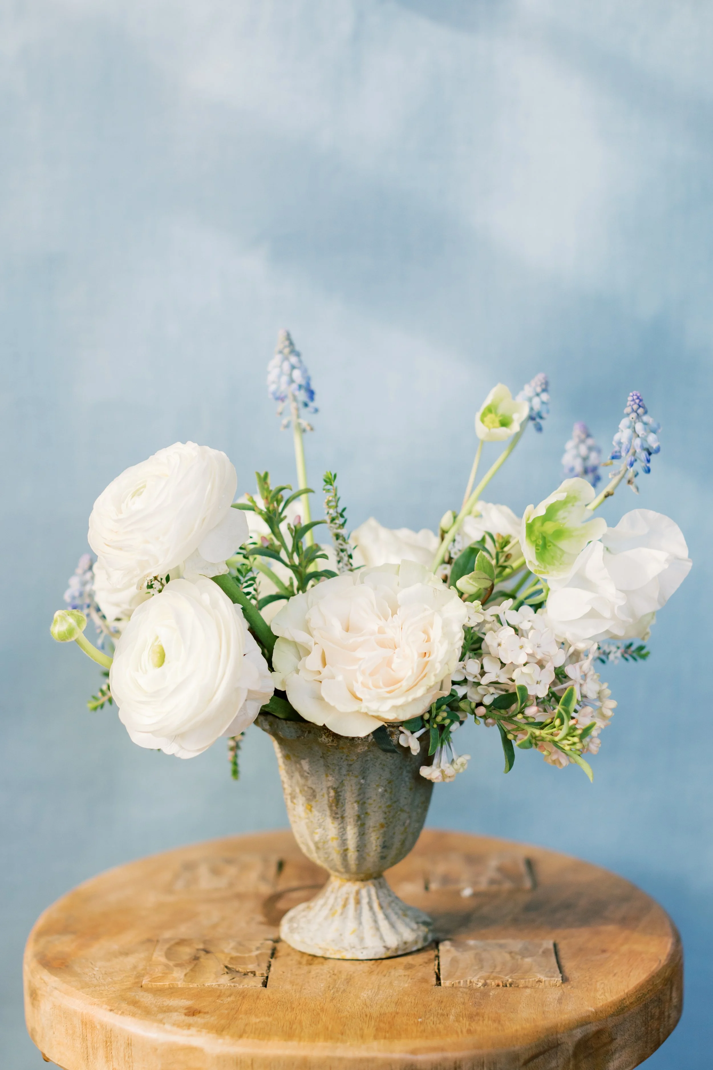 A floral arrangement with white and light pink flowers in a rustic vase on a wooden table against a soft blue background.