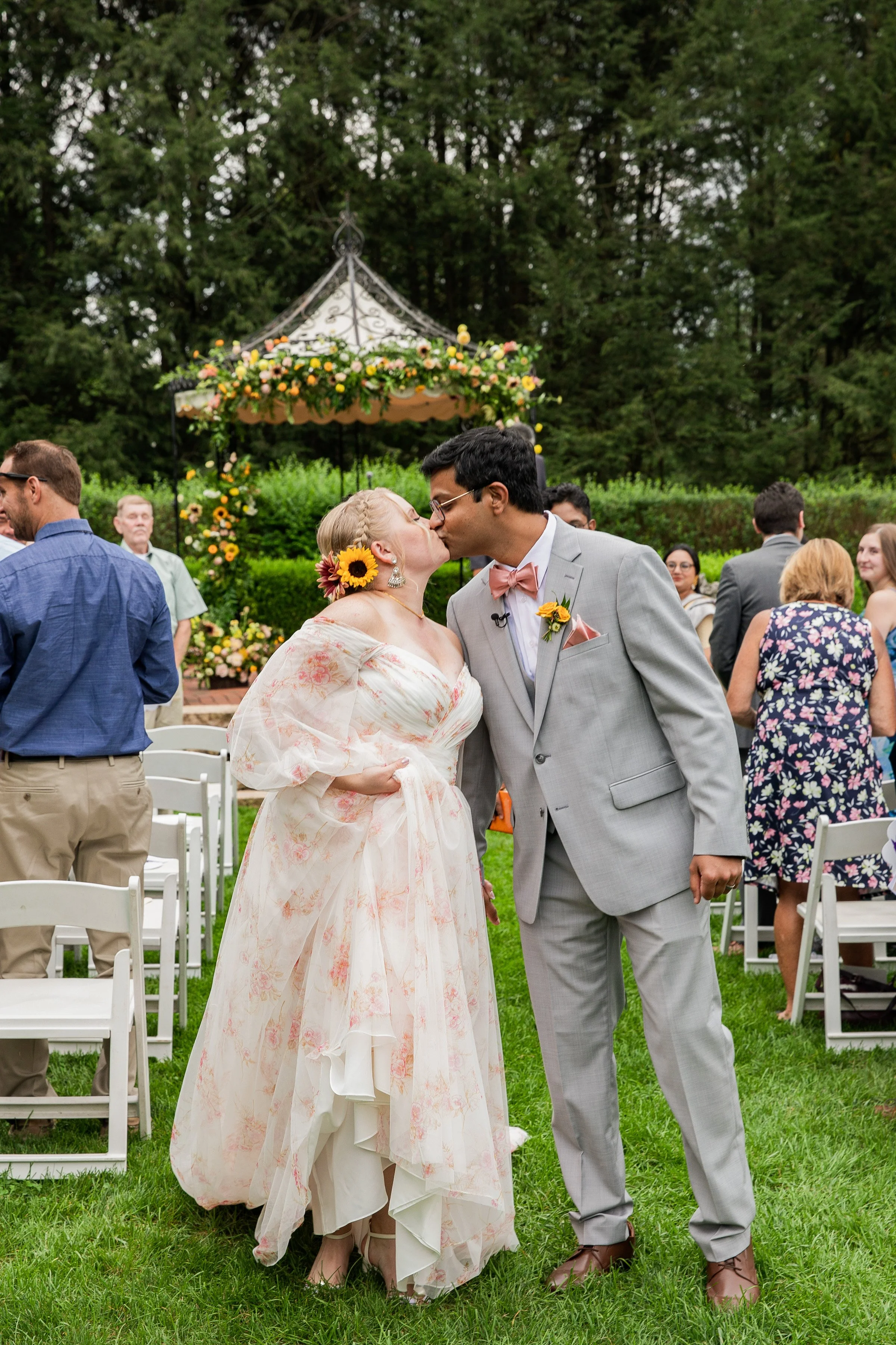 A bride and groom kissing at an outdoor wedding ceremony, surrounded by guests. The bride wears a floral dress and sunflower accessories, while the groom is in a gray suit with a pink bow tie. A floral arch is in the background with greenery around.