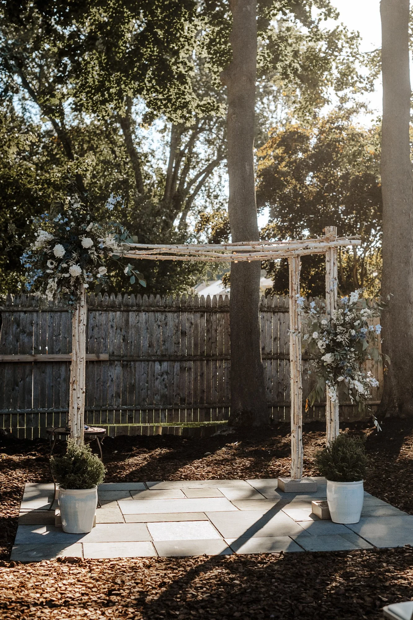 Forest-inspired birch-pole wedding arbor with asymmetrical white and blue floral accents and potted boxwoods on a stone patio