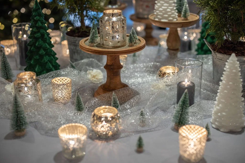 Candlelit winter tablescape with mercury glass votives, ceramic and bottle-brush trees on wooden risers, sparkly tulle and potted evergreens at The Inn at Longshore in Westport, Connecticut