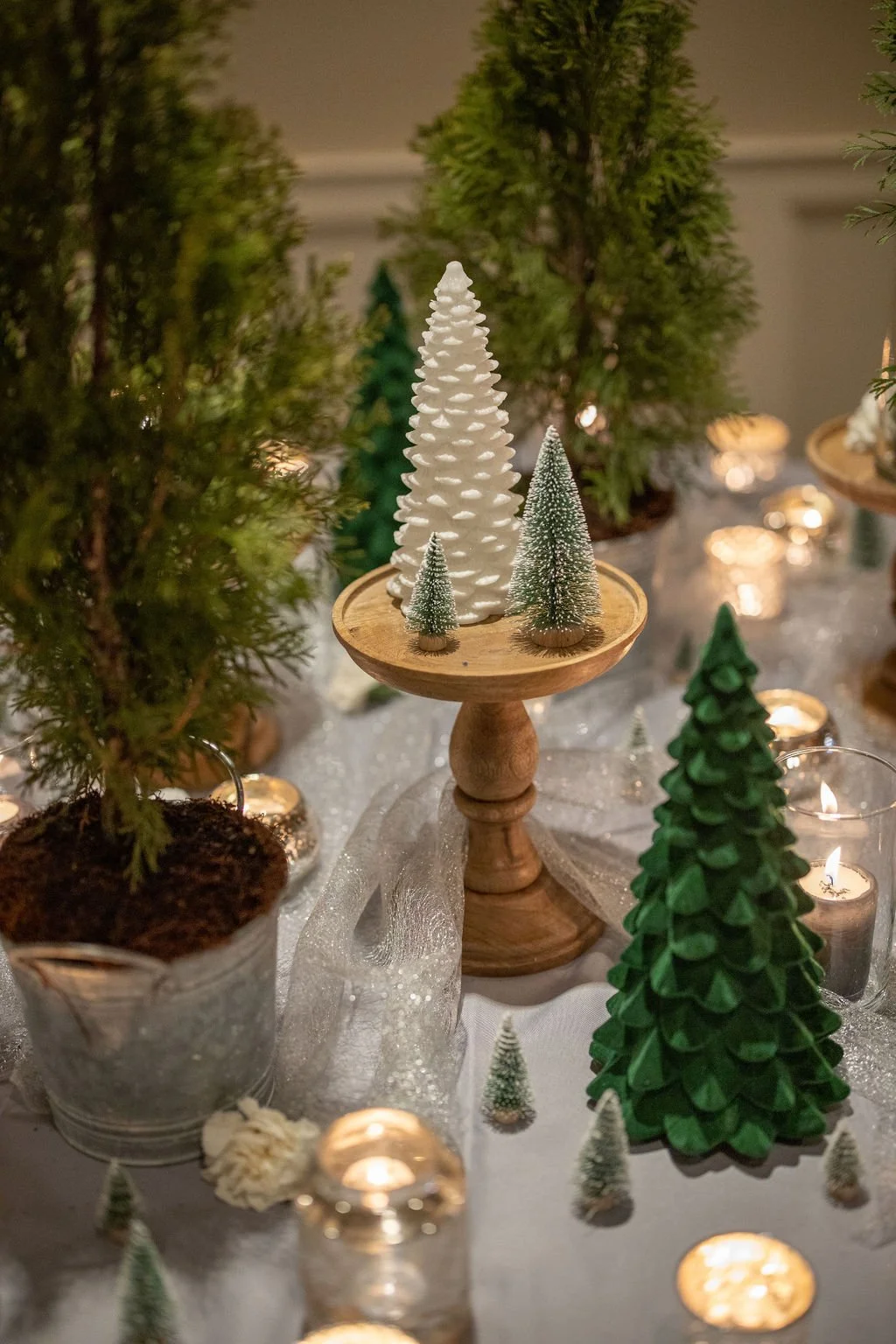 Winter tablescape with potted evergreen trees in galvanized pails, ceramic and bottle-brush trees on a wooden riser, sparkly tulle and mercury glass votives