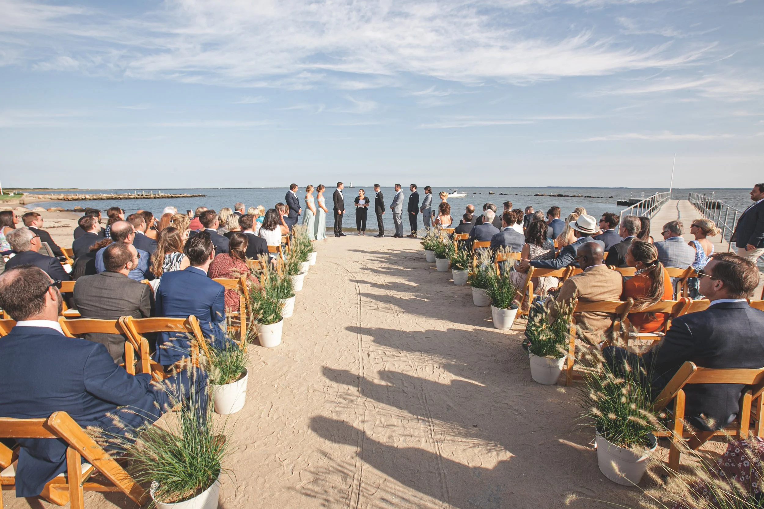 Wooden ceremony chairs face the water's edge at a private Saybrook Point residence, framed by beach grass and open sky.