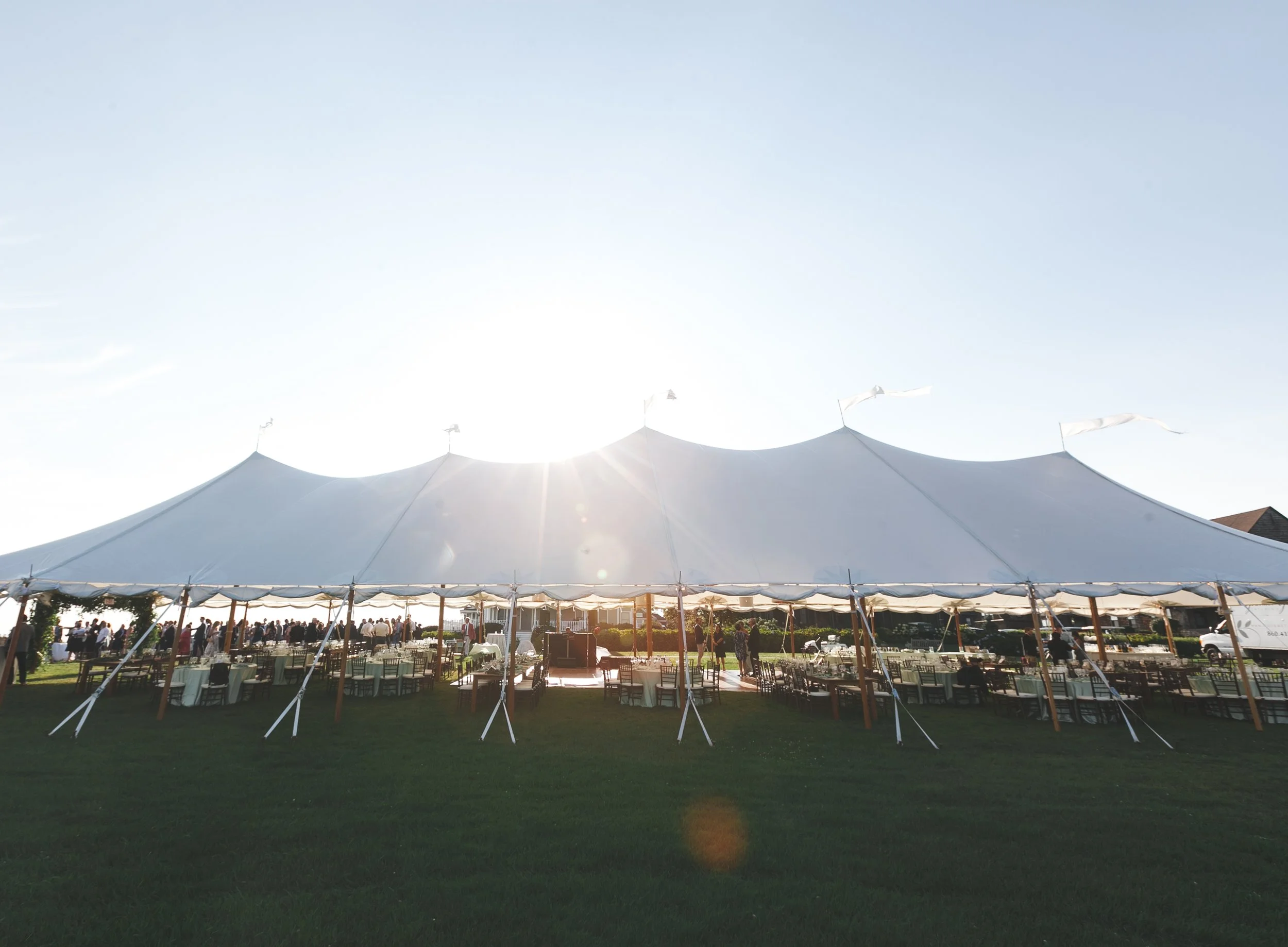 Wooden tent poles rise beneath draped sailcloth, framing a coastal Connecticut reception at golden hour.