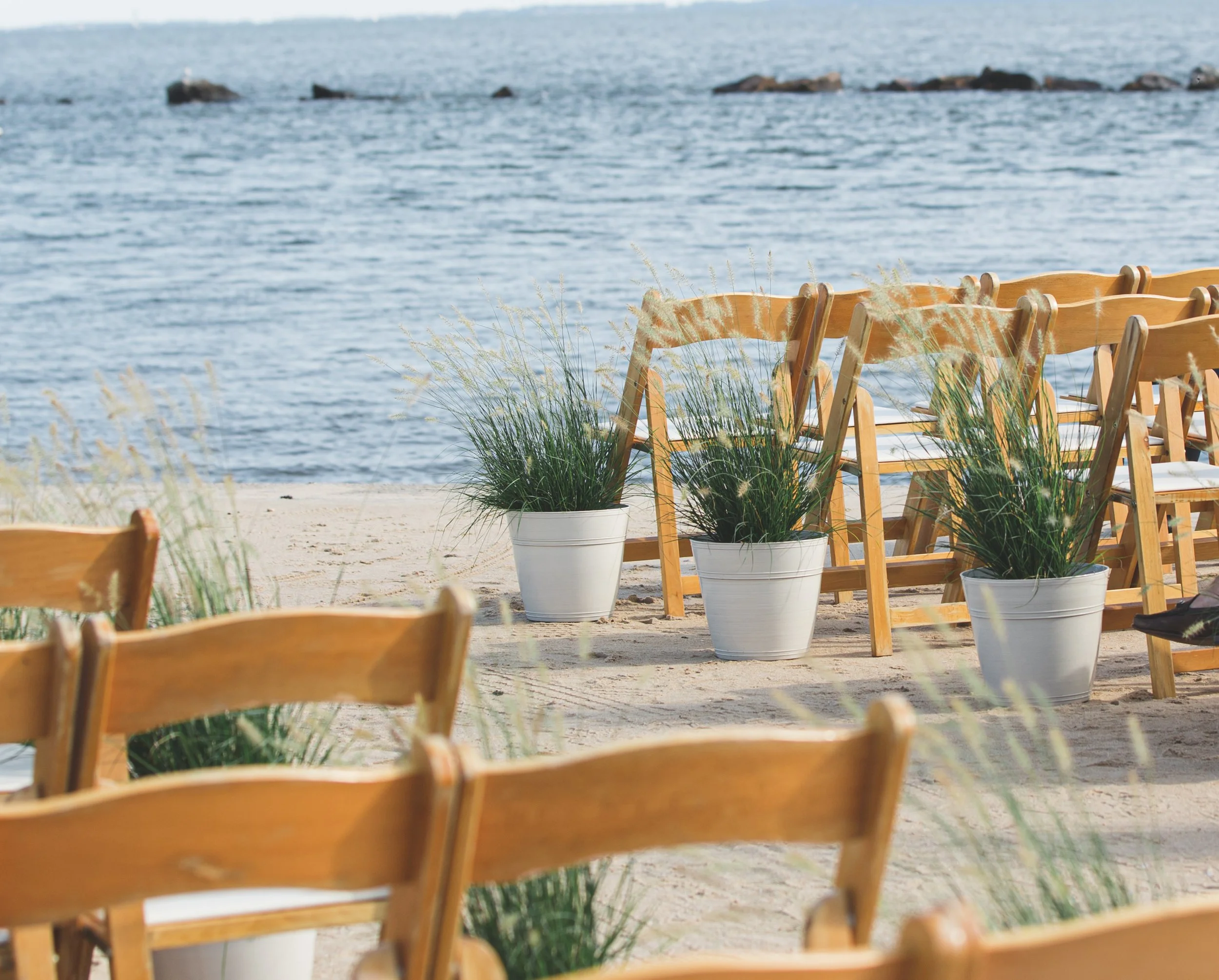 Wooden folding chairs line a sandy beach ceremony aisle, framed by potted ornamental grasses at a Saybrook Point wedding by First Blush.