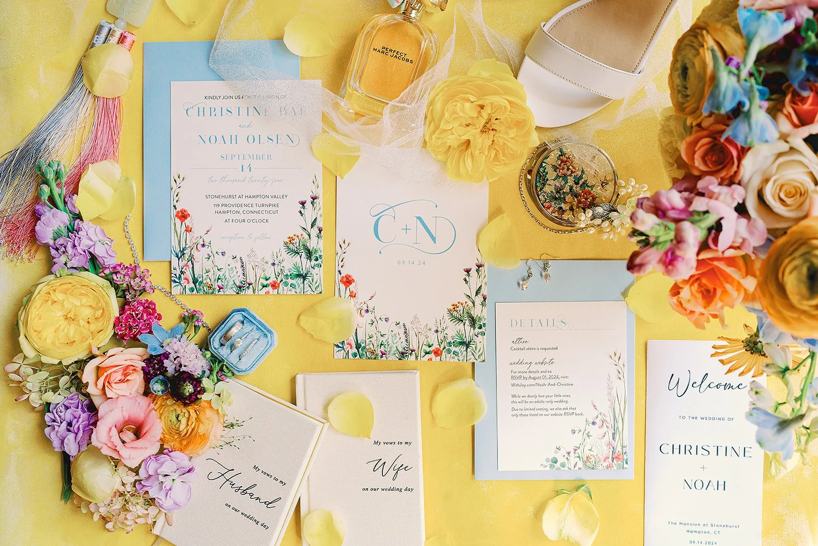 Flat lay of colorful wedding stationery and accessories including invitations, floral arrangements, jewelry, perfume, and a bridal shoe on a yellow background.