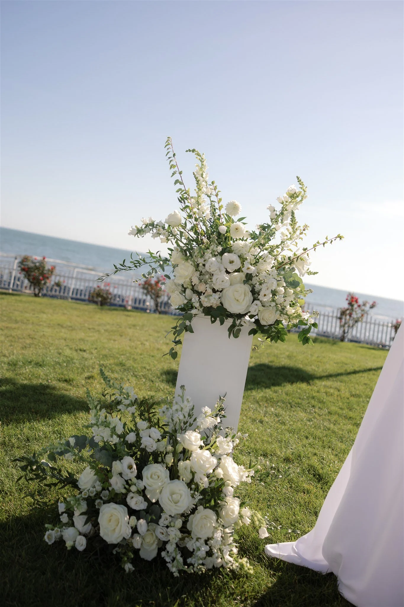 Aisle view of an all-white waterfront ceremony framed by airy florals and a hazy blue summer sky.