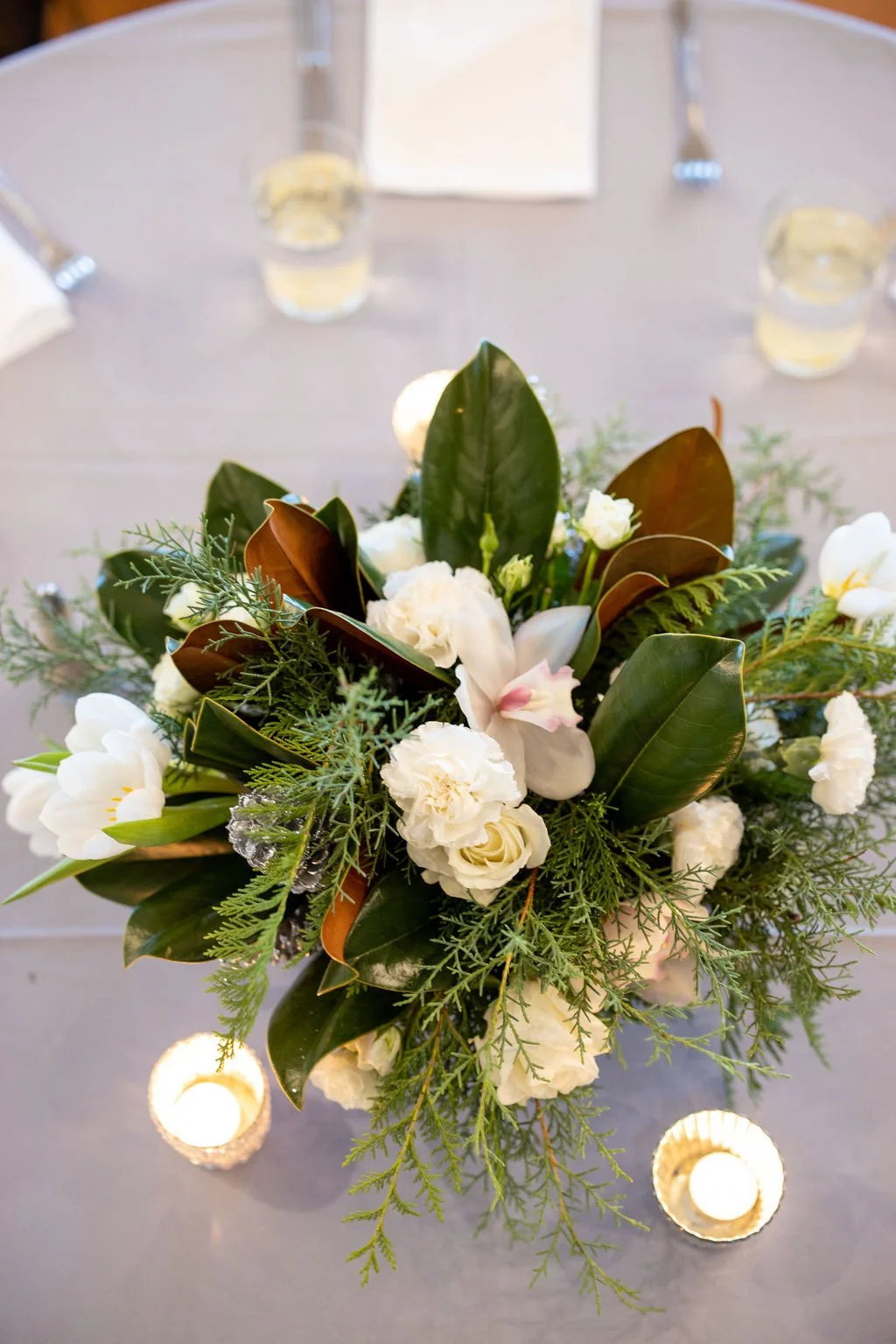 Overhead view of a low centerpiece with magnolia leaves, cedar, white roses, carnations, tulips and an orchid between two mercury glass votives