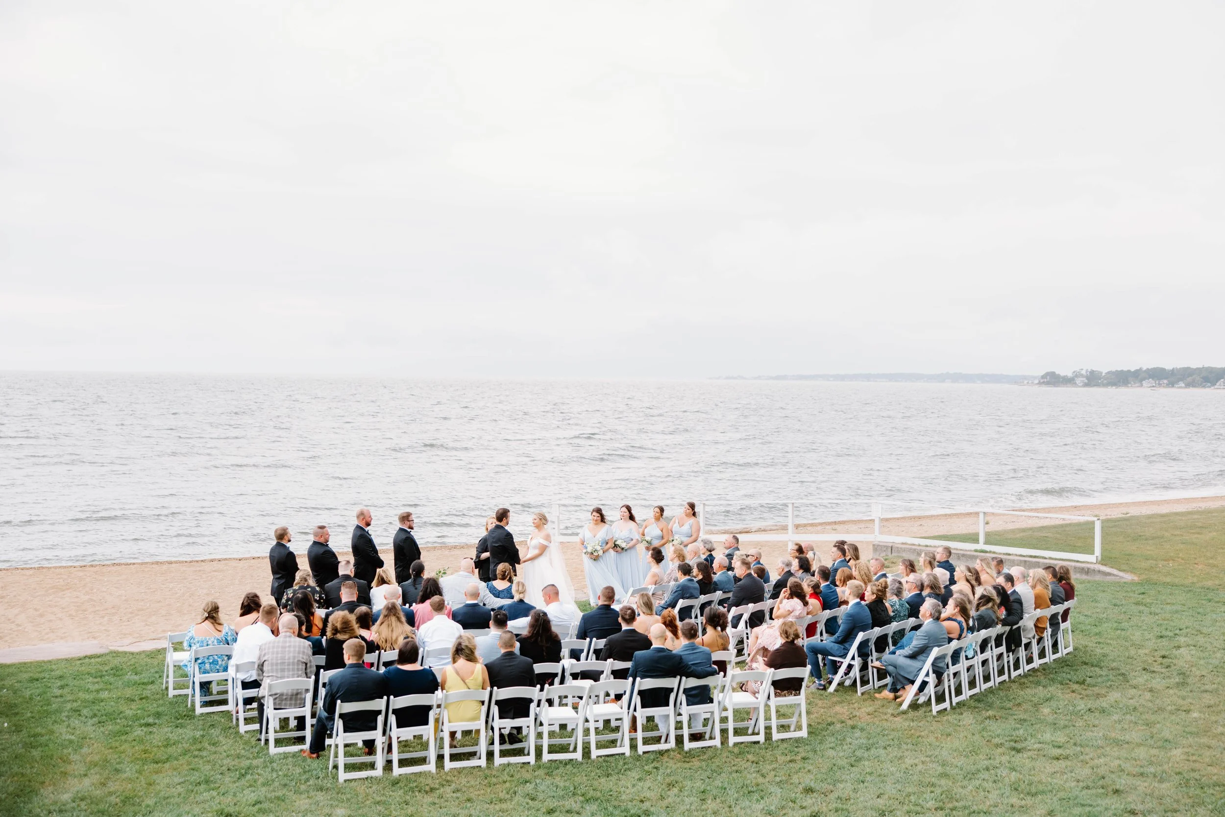 Wedding party in dusty-blue dresses and black tuxes posing by the Connecticut shoreline.