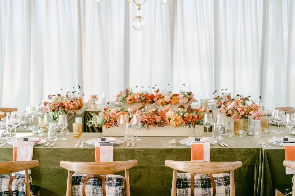 Elegant dining table set with floral centerpieces, glassware, and place settings with napkins. Wooden chairs have plaid blankets draped over them. The setting is in a well-lit room with white curtains.