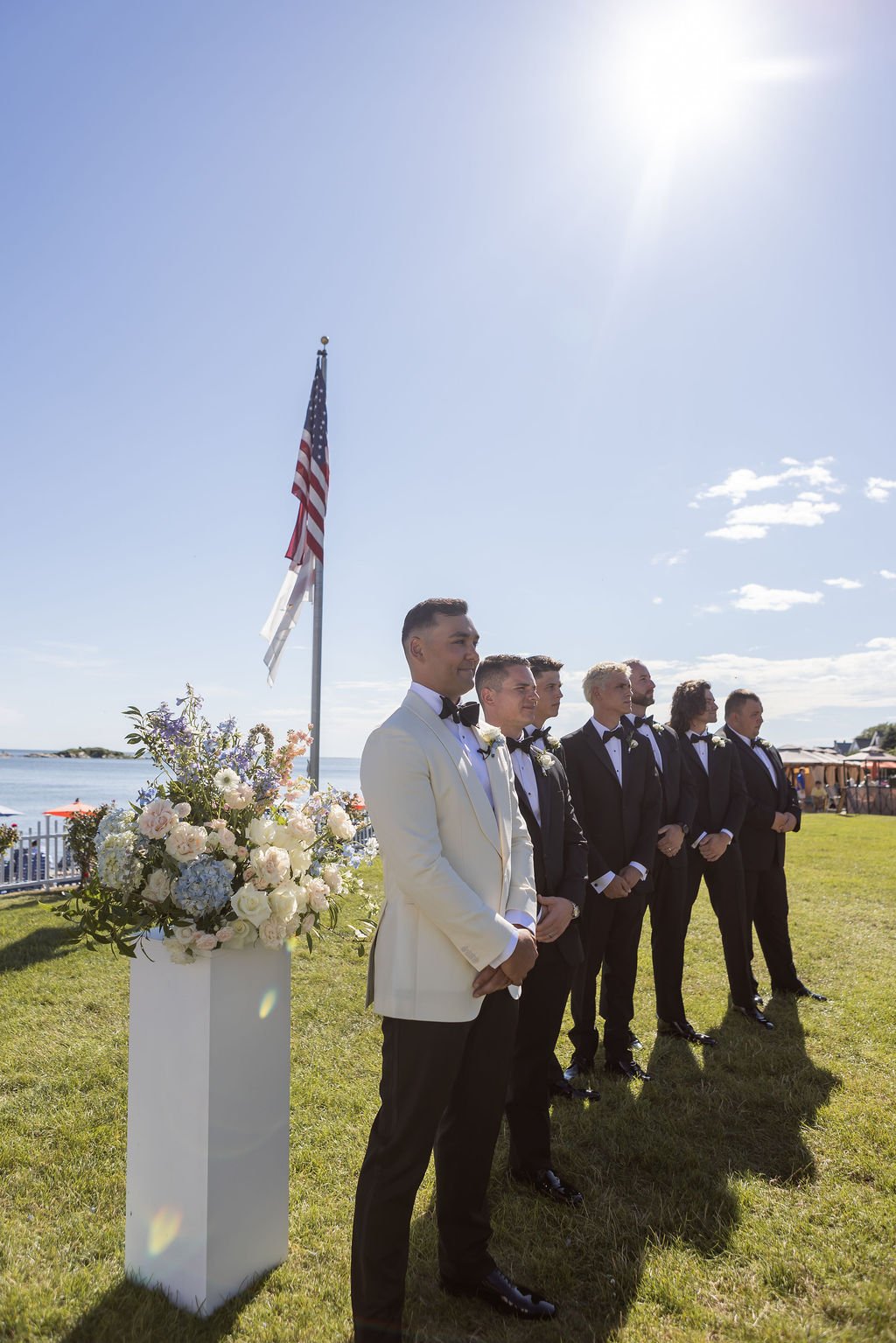 White column topped with a garden-style floral arrangement of white hydrangea, blue delphinium, and local foliage at a waterfront ceremony at Water's Edge Resort Connecticut, First Blush Floral Design