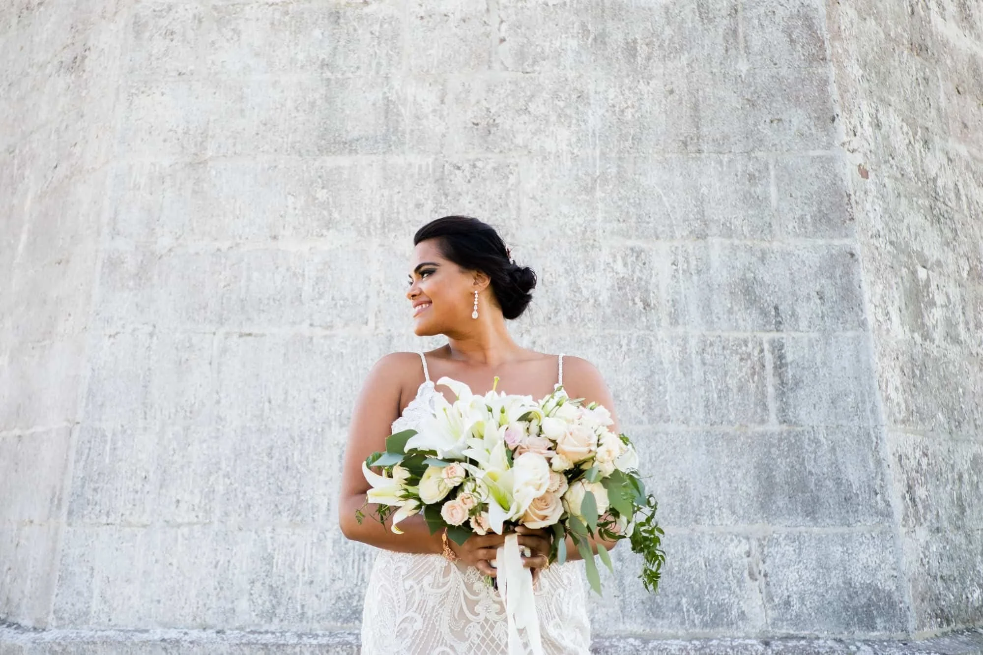 Smiling bride holding a white lily bouquet with eucalyptus and a flowing ivory ribbon in front of a weathered stone wall