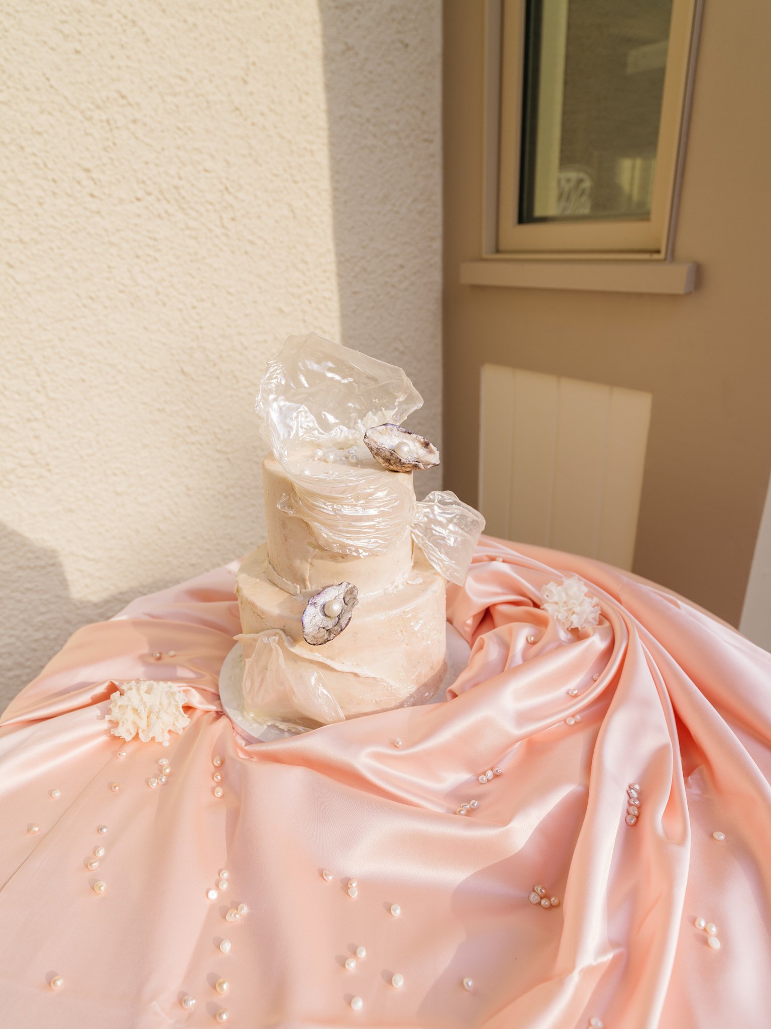 Two-tier cake by Andy Cakes NYC wrapped in cellophane and adorned with oyster shells and pearls on blush satin linen, coastal gilded age bridal shower design