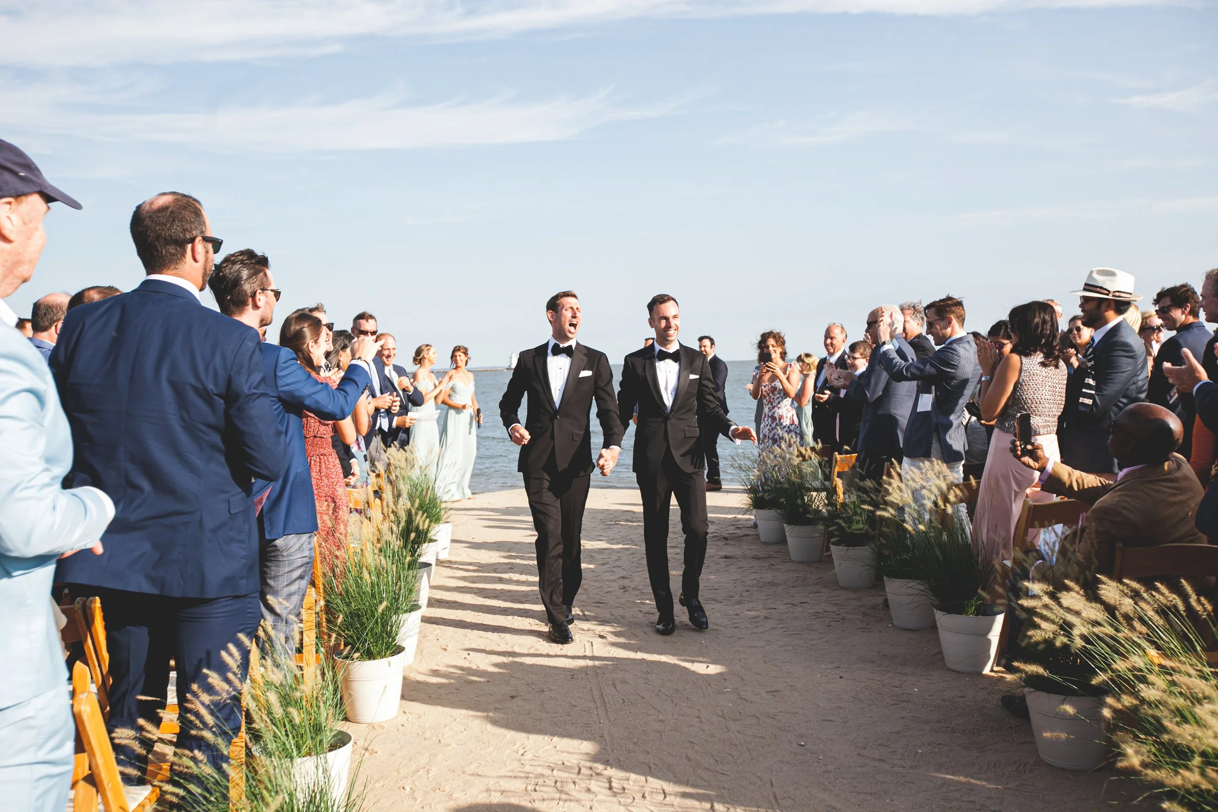 Two grooms in tuxedos walk hand in hand along a dock, silhouetted against a setting sun on the Connecticut shoreline.