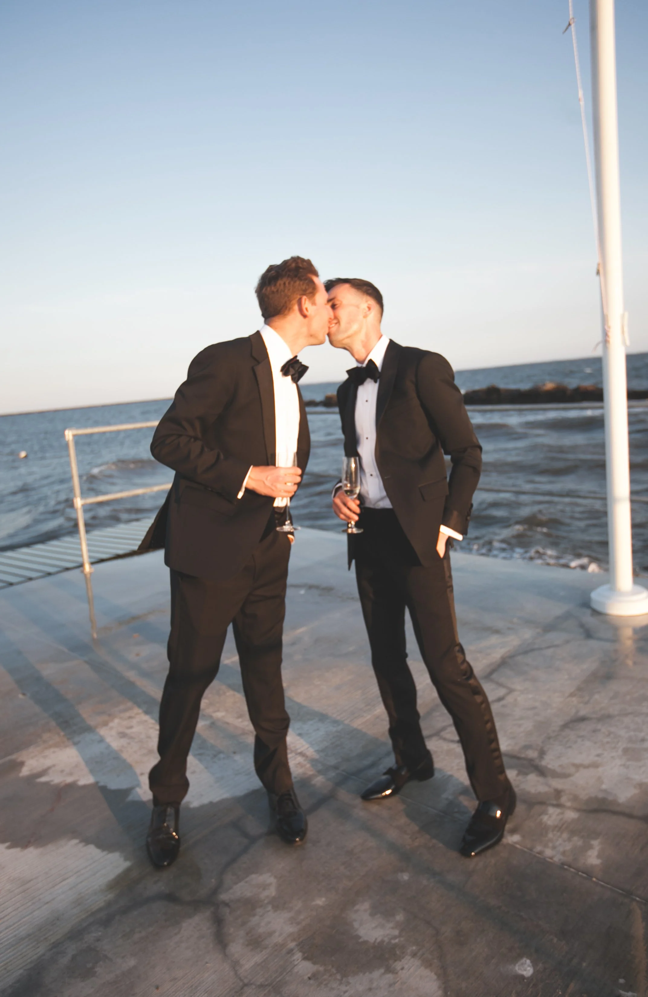 Two grooms hold champagne glasses on a waterfront dock, with shoreline homes visible across the water at Saybrook Point.