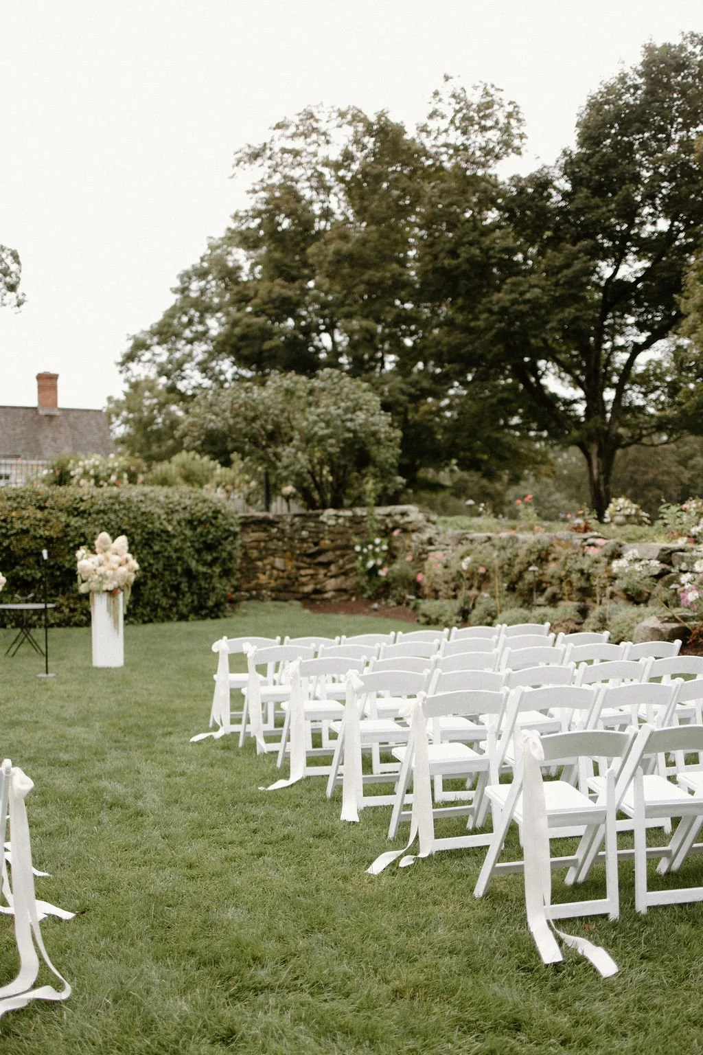 Outdoor wedding ceremony setting with white guest chairs arranged along a tree-lined garden aisle.