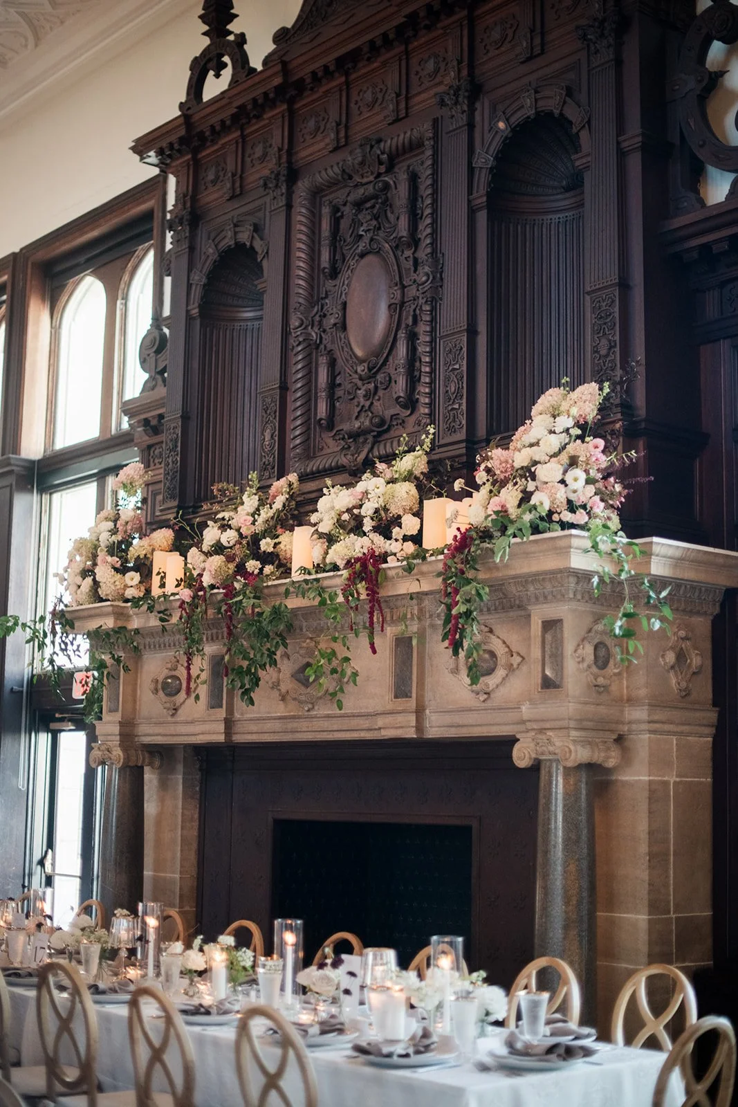 Elegant indoor dining area with a long table set for a meal, featuring candles, white flowers, and glassware. A large ornate wooden fireplace mantel decorated with pink and white floral arrangements and greenery is in the background, with intricate carvings. Natural light filters in through tall windows.