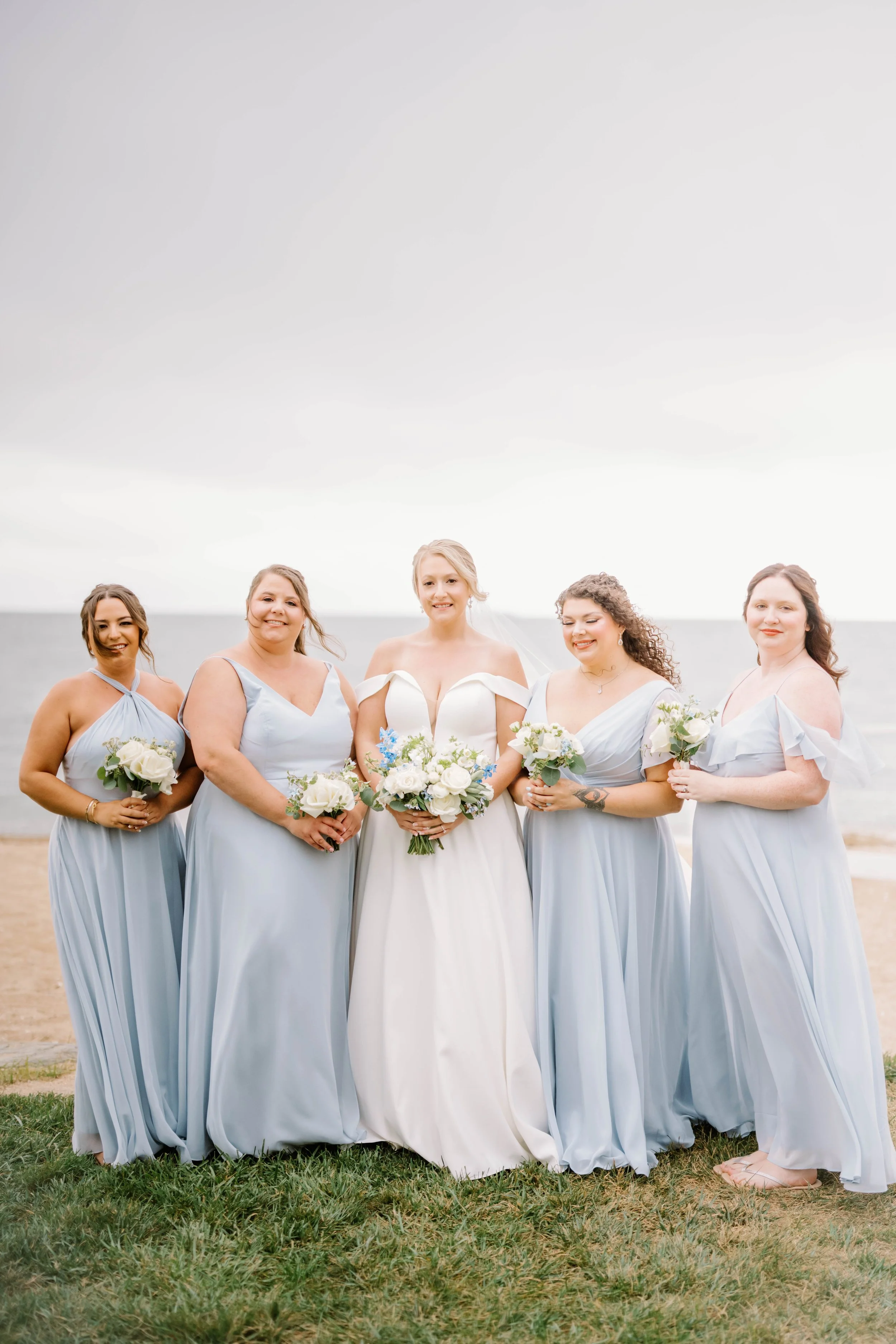 Bridesmaids lined up in dusty-blue gowns holding small posy bouquets of white and blue flowers.