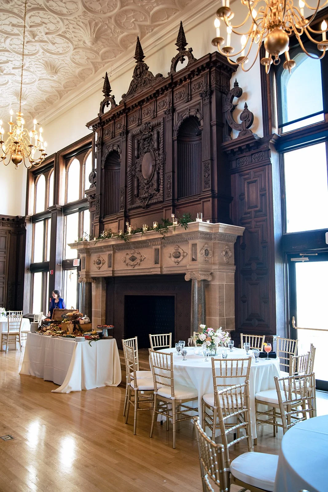 Branford House reception hall in Groton, Connecticut, with a carved dark wood overmantel, a stone fireplace with candlelit greenery garland, a grazing table, gold chiavari chairs and a blush floral centerpiece