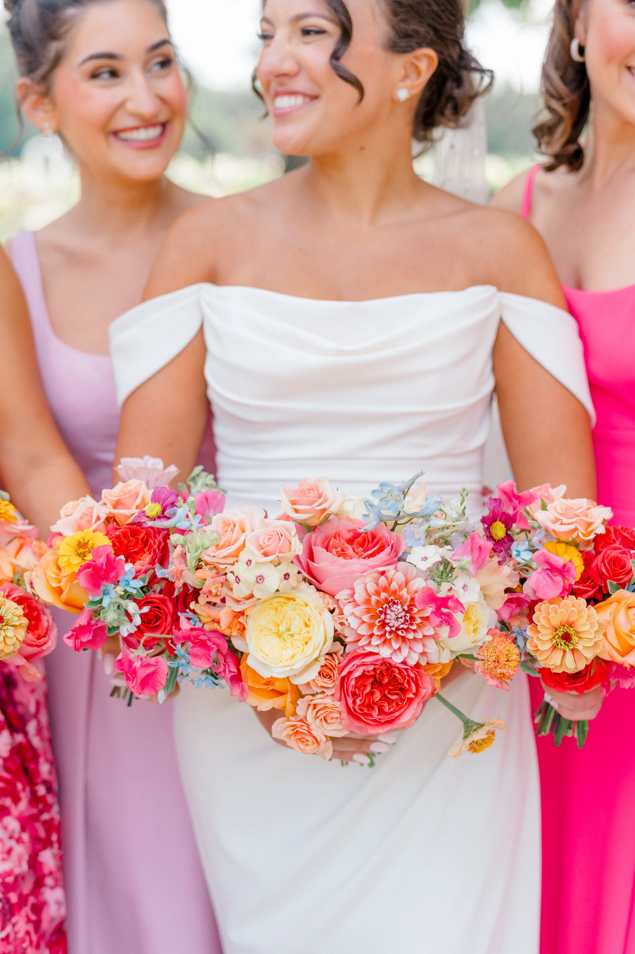 Bridesmaid bouquets in peach, coral, and yellow with accents of blue delphinium and zinnias.