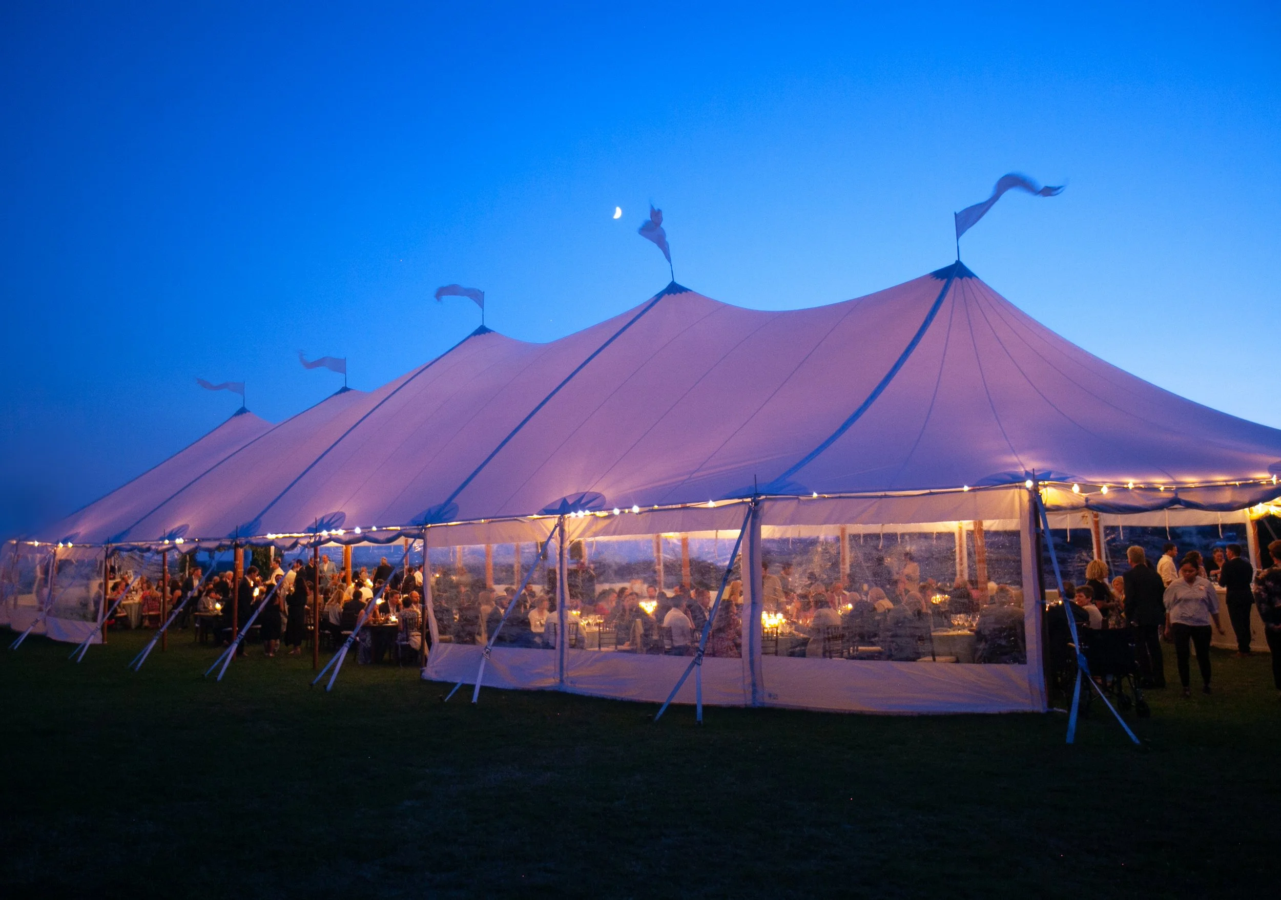 Wooden tent poles rise beneath draped sailcloth, framing a coastal Connecticut reception at golden hour.