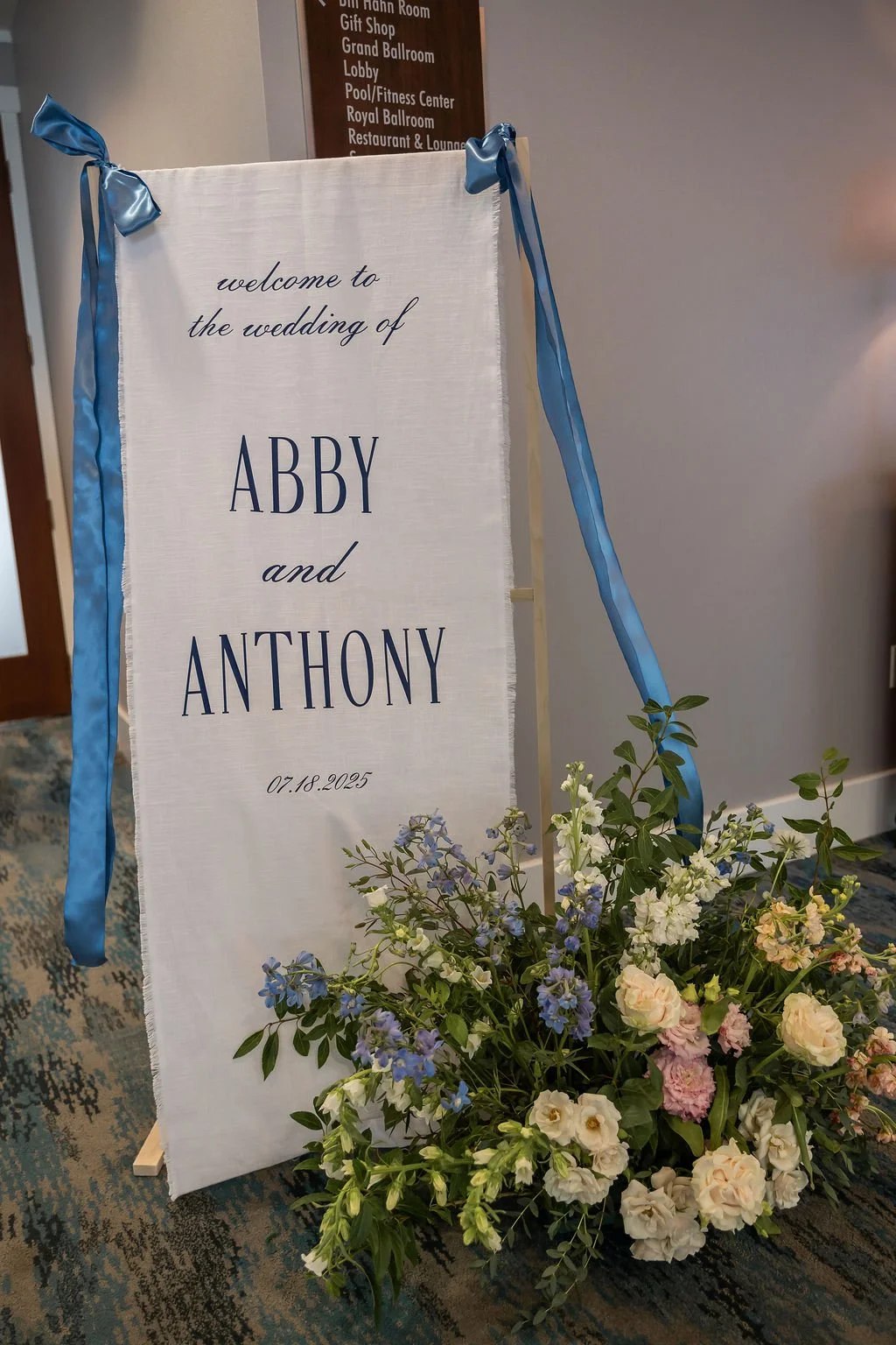 Low garden-style wedding centerpiece in a glass compote vessel with white roses, blush blooms, and blue delphinium on a white linen reception table at Water's Edge Resort Connecticut, florals by First Blush Floral Design