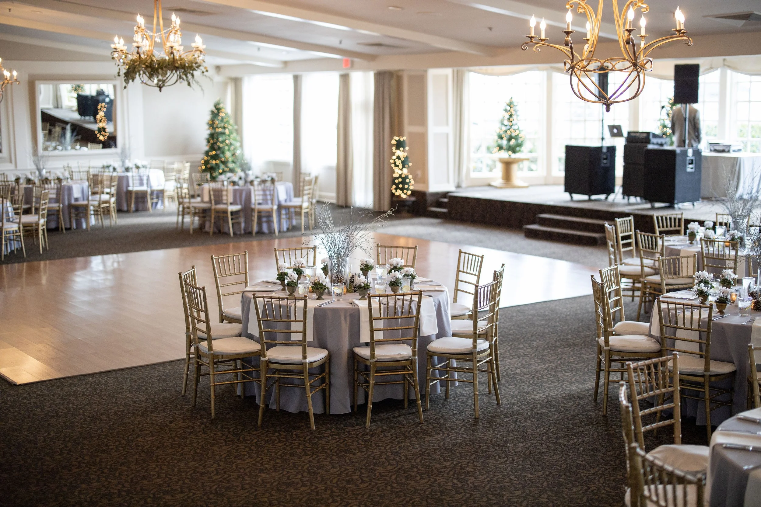 Ballroom at The Inn at Longshore in Westport, Connecticut, set for a winter corporate event with gray linens, gold chiavari chairs, glittered branch centerpieces and lit Christmas trees