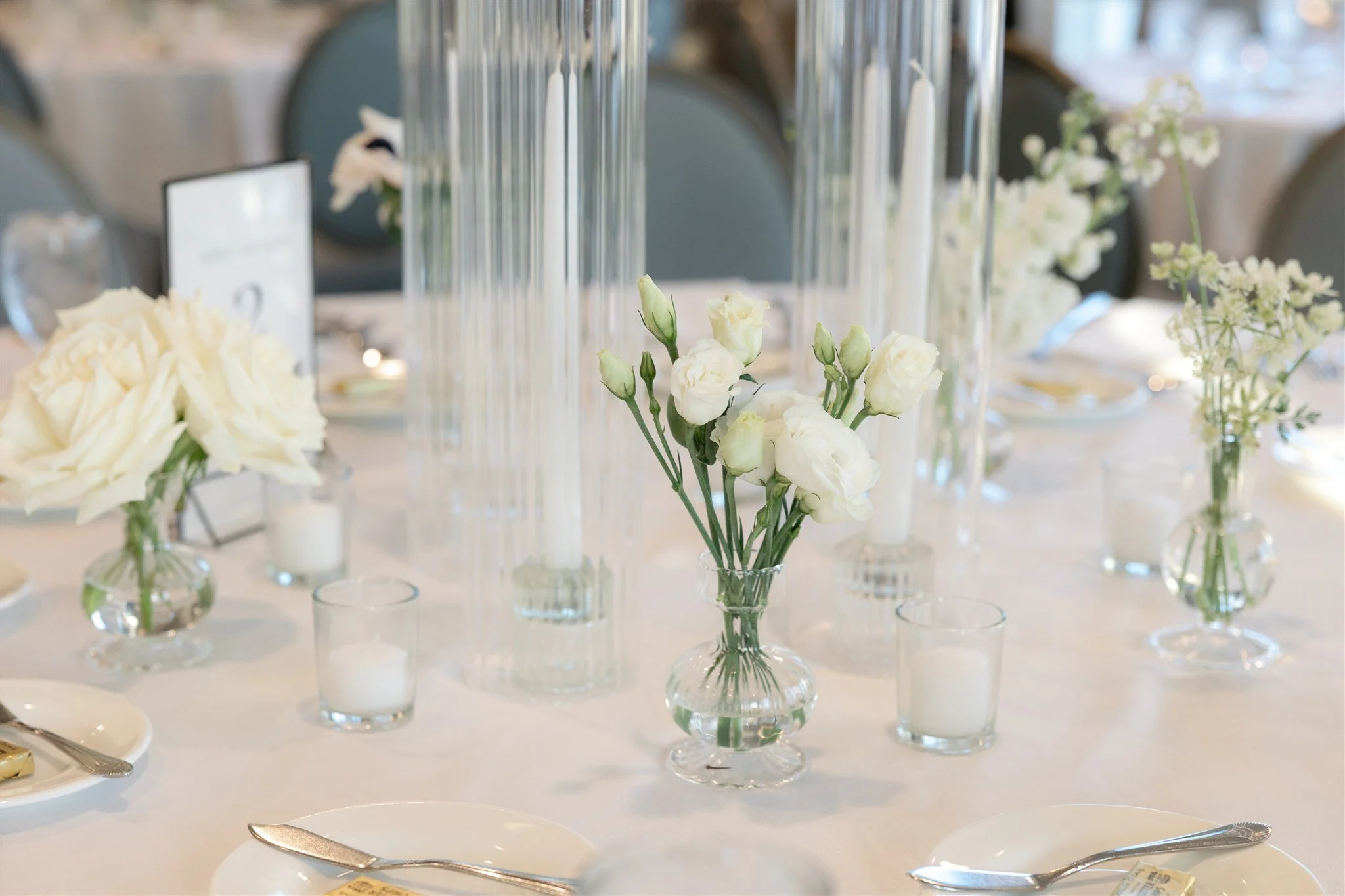 Candlelit Water’s Edge reception with round tables styled in white linens, glass hurricanes, and delicate all-white budvase florals.