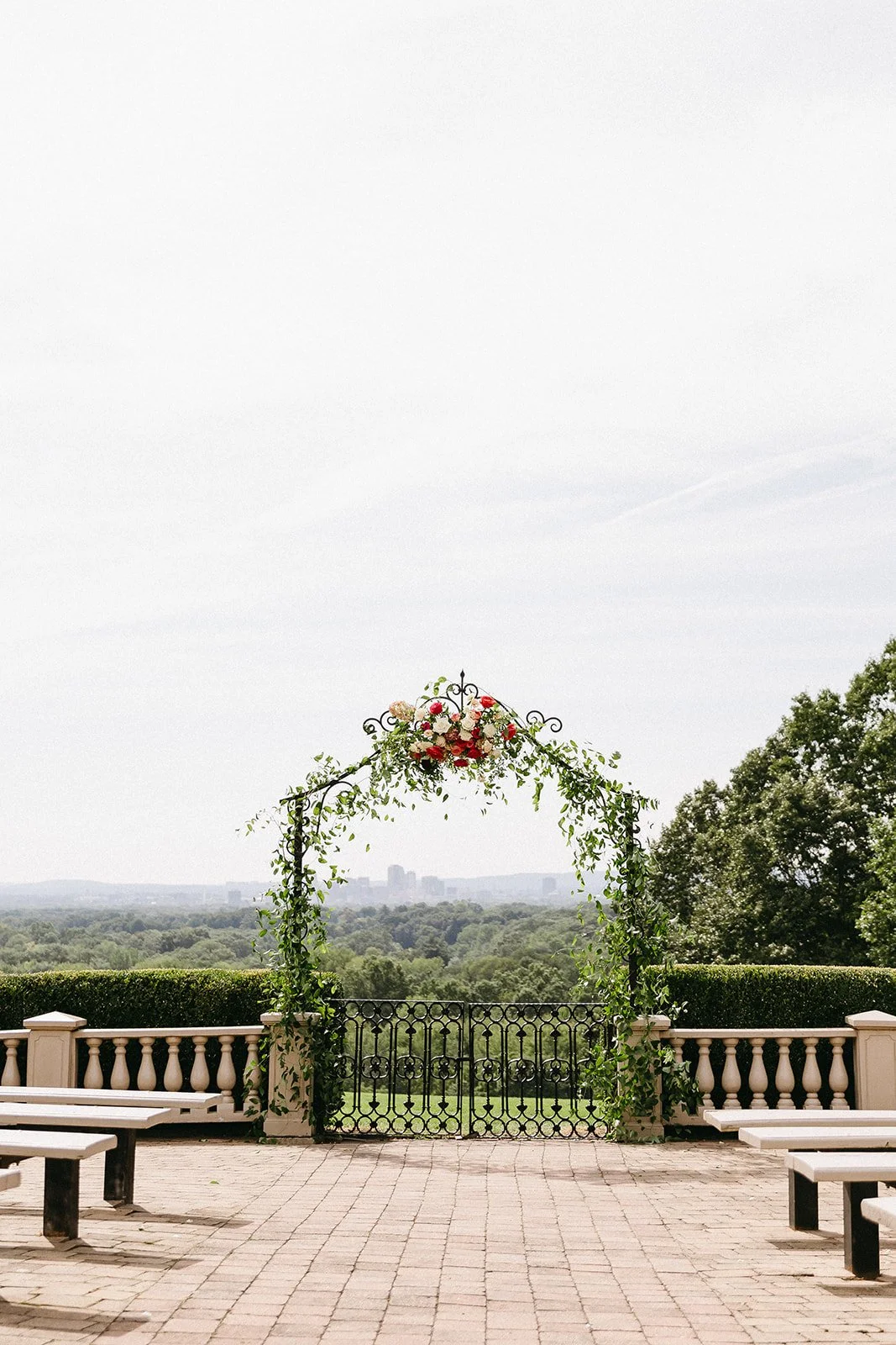 Iron ceremony arch draped in greenery with a cluster of blush, pink and cherry red roses, overlooking the Hartford skyline at Wickham Park in Manchester, Connecticut