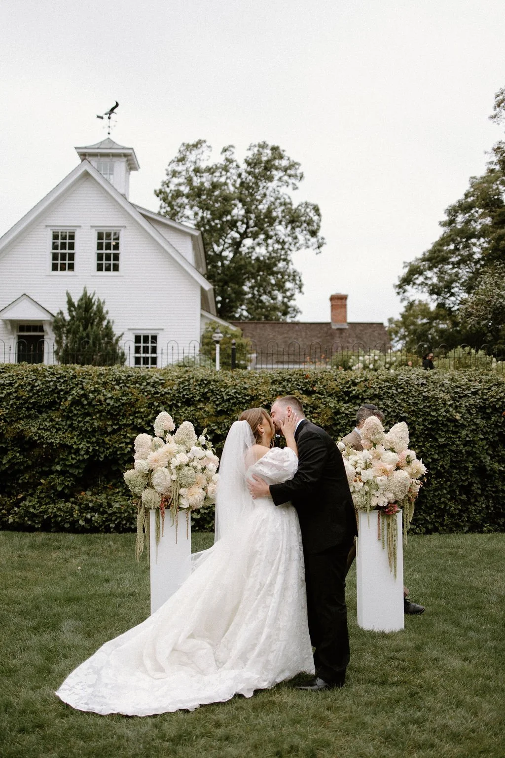 Bride and groom kissing between two white ceremony columns topped with lush ivory and blush garden flowers at Smith Farm Gardens in Connecticut.