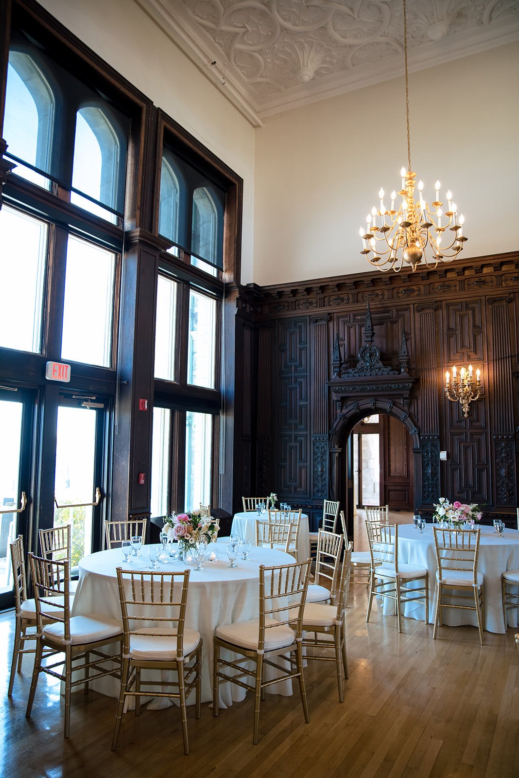 Branford House reception room with a brass chandelier, arched windows and dark wood paneling, styled with soft blush wedding flowers