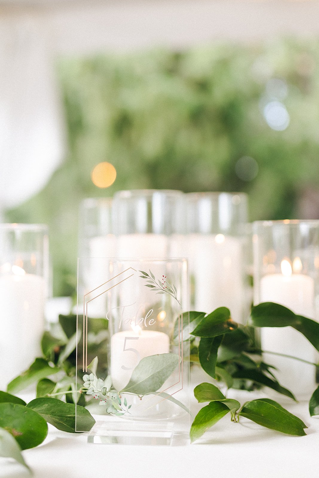 Close-up of white pillar candles in clear glass hurricanes with loose greenery arranged along a garden wedding reception table.