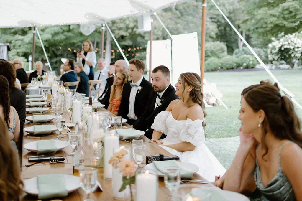 Bride and groom seated at a candlelit reception table with garden-style flowers beneath a sailcloth tent.