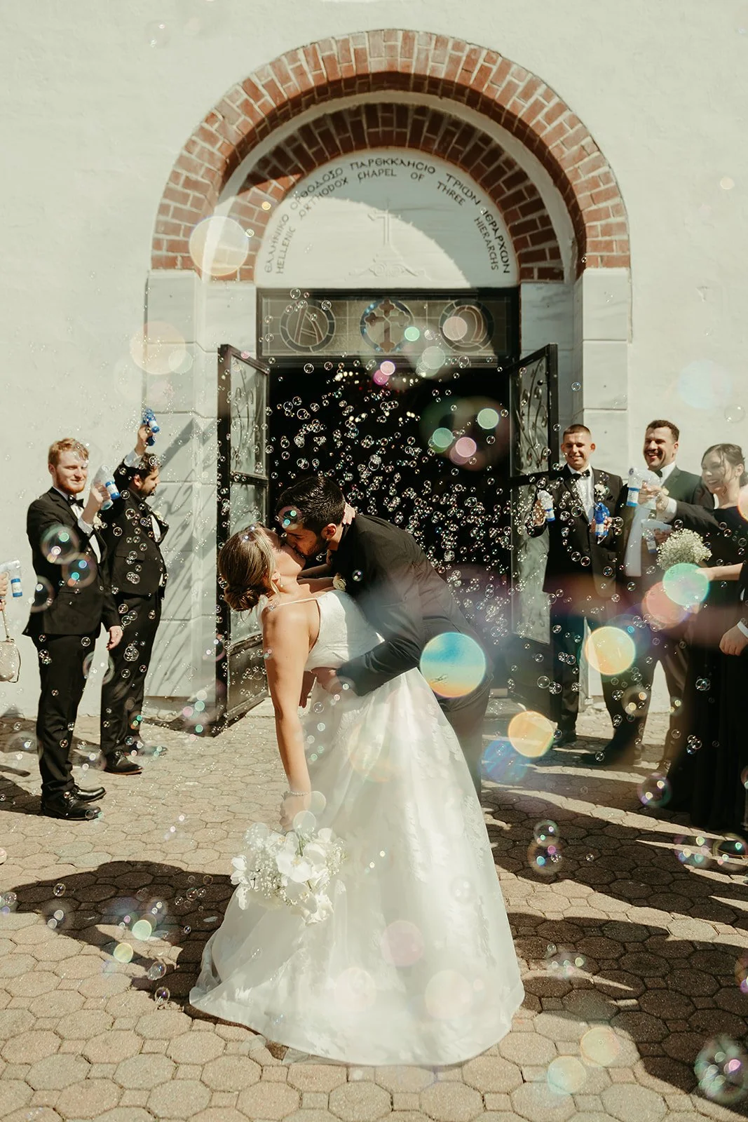 Bride and groom kissing outside a chapel surrounded by bubbles, with guests in suits and a dress cheering and blowing bubbles.
