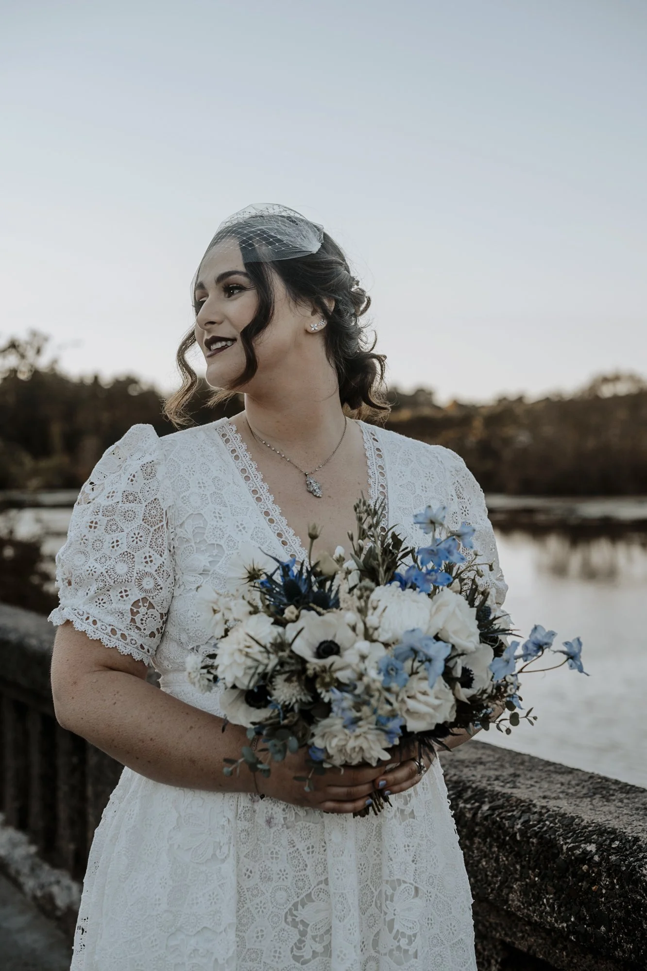 Bride in a lace dress and birdcage veil holding a white and blue bouquet with delphinium and thistle, standing by lake.