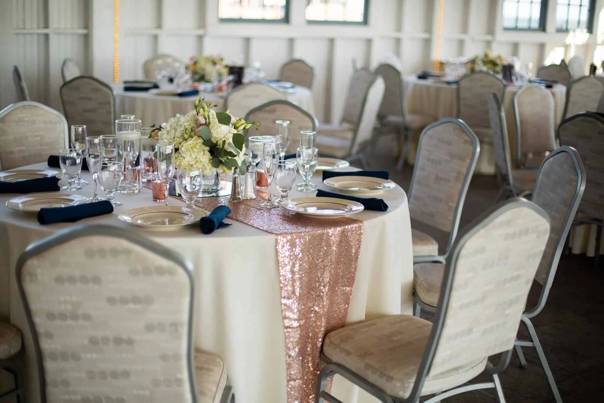 Reception table with a white hydrangea centerpiece in clear glass, navy napkins and a rose gold sequin runner inside the Lighthouse Point carousel pavilion