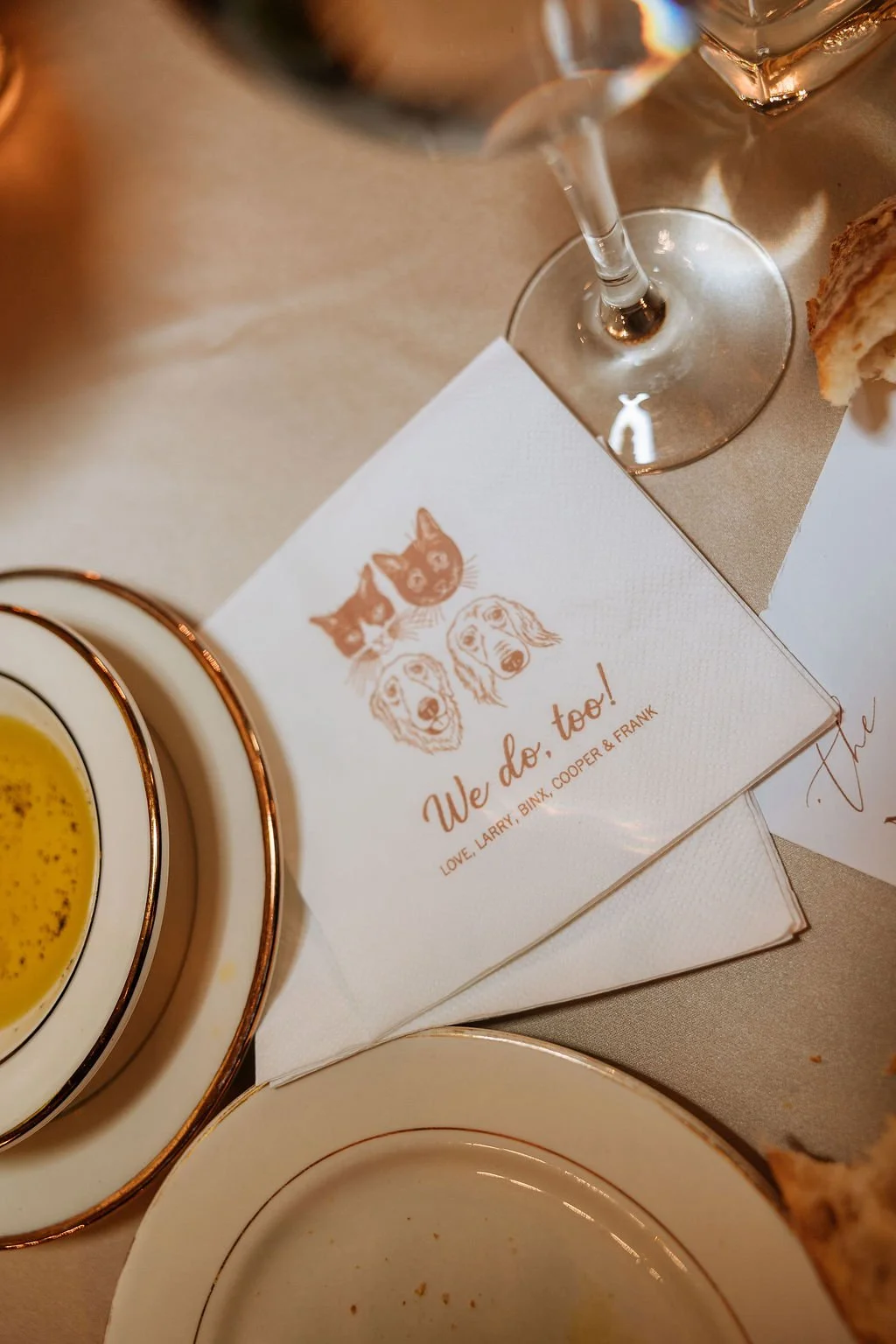 Close-up of a personalized white wedding napkin printed with a warm-toned floral illustration and the words 'We do, too!' beside gold-rimmed plates and glassware at a Waveny House reception.
