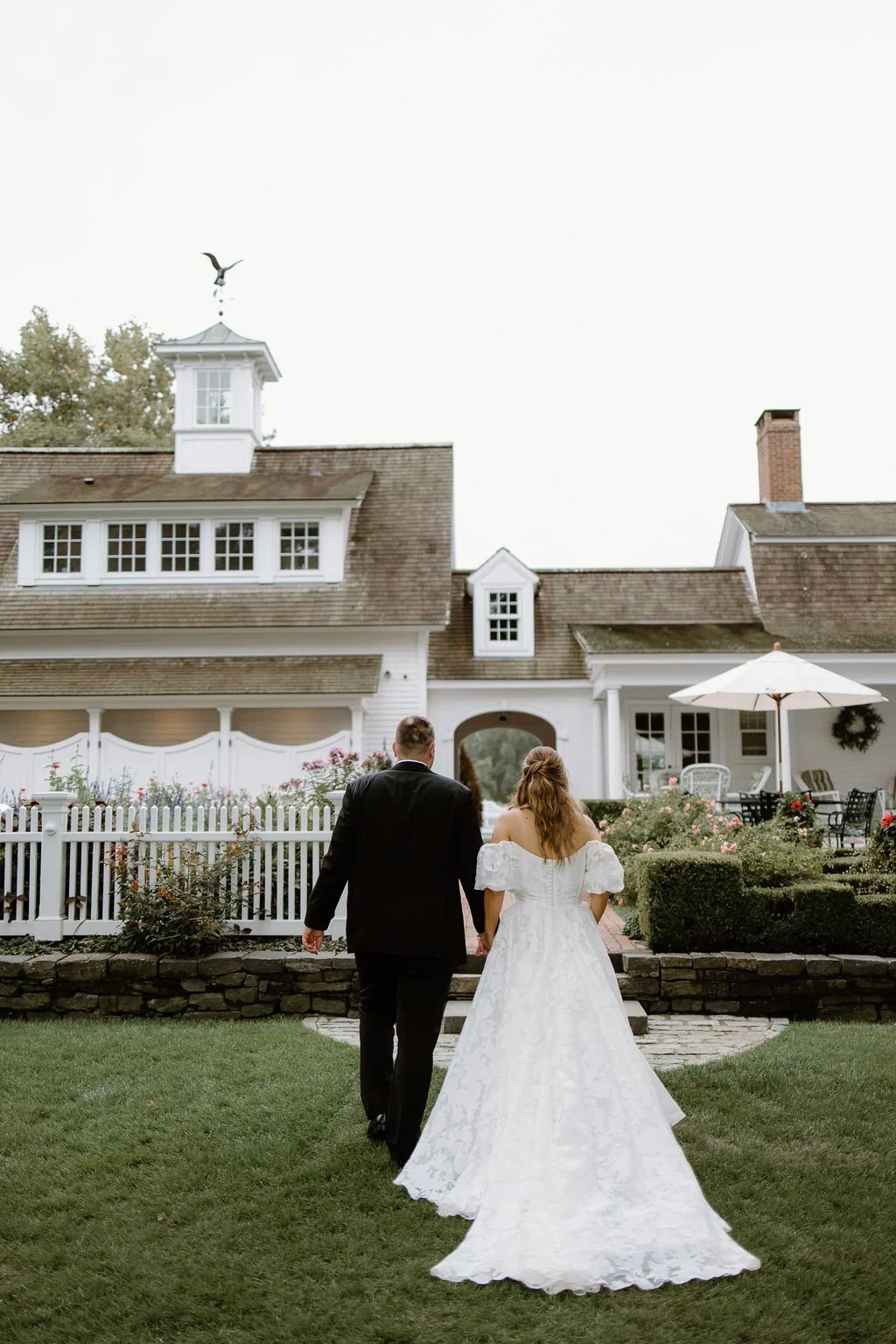 Bride and groom walking together across the lawn at Smith Farm Gardens, with the historic white estate and lush greenery in the background.