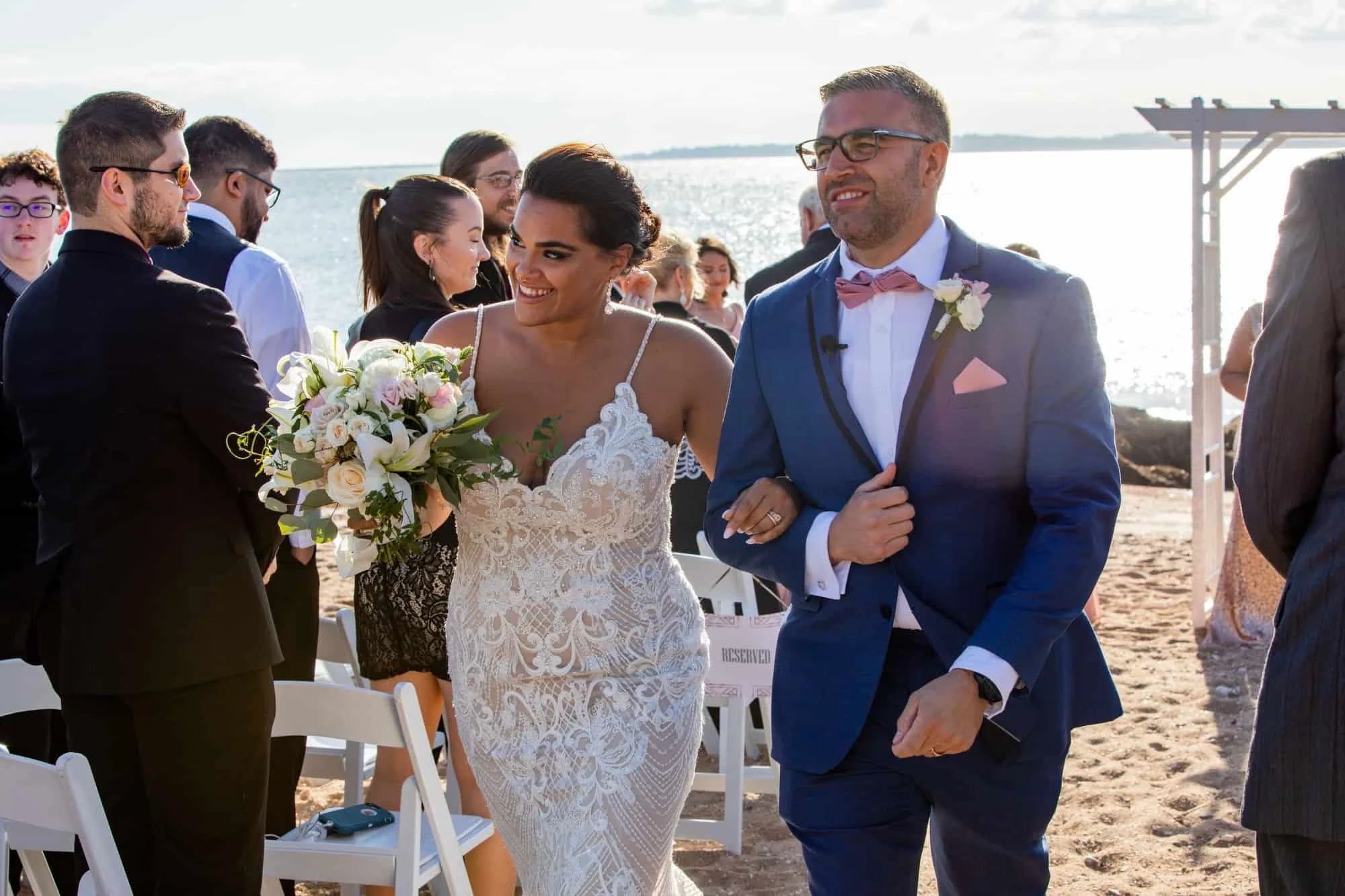 Bride in a lace gown holding a cascading bouquet of white lilies, lisianthus and spray roses against the stone lighthouse wall at Lighthouse Point Park, New Haven, CT