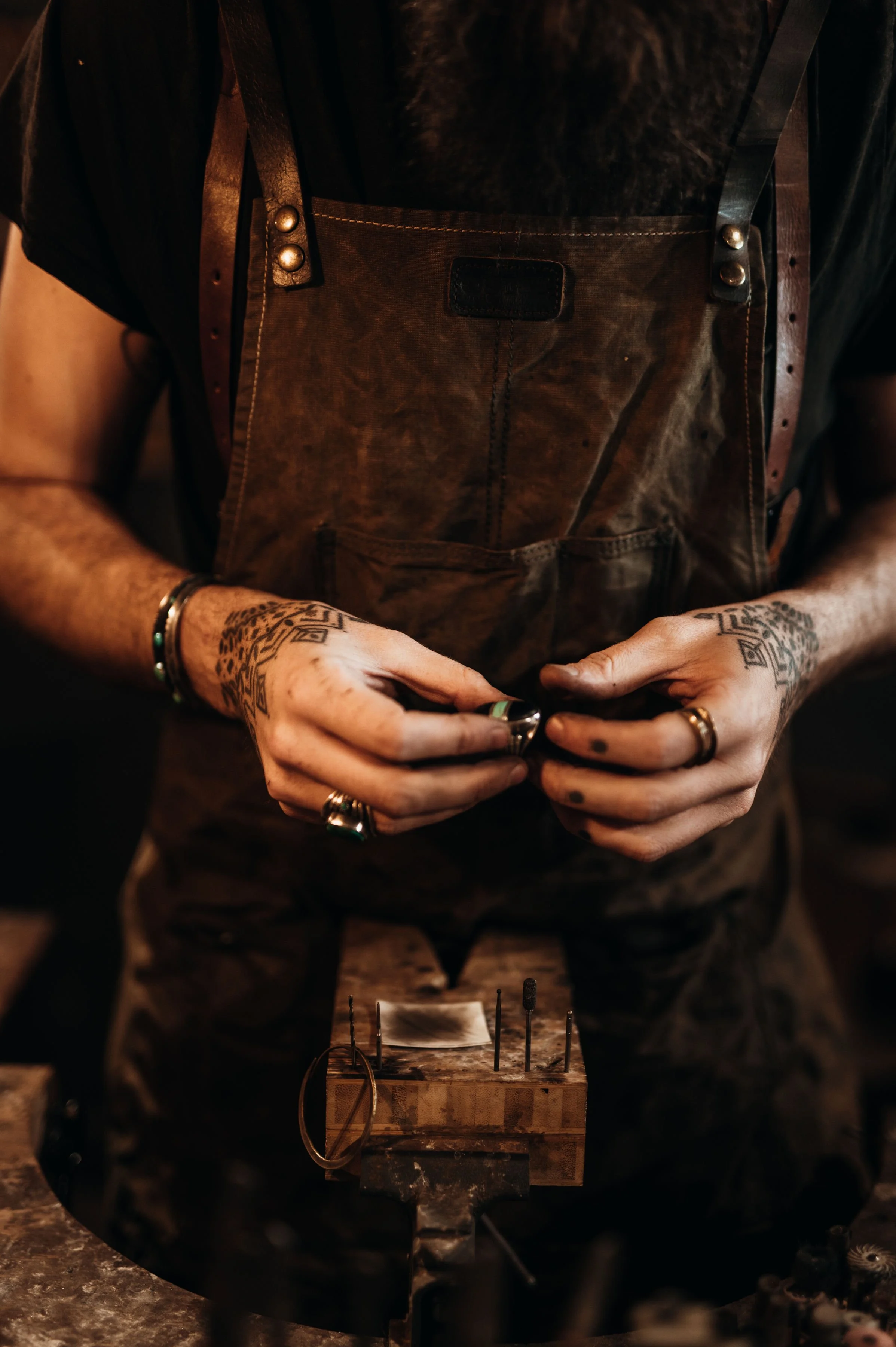 Close-up of a person wearing a brown apron with tattooed hands adjusting a small object, standing at a workbench with tools.
