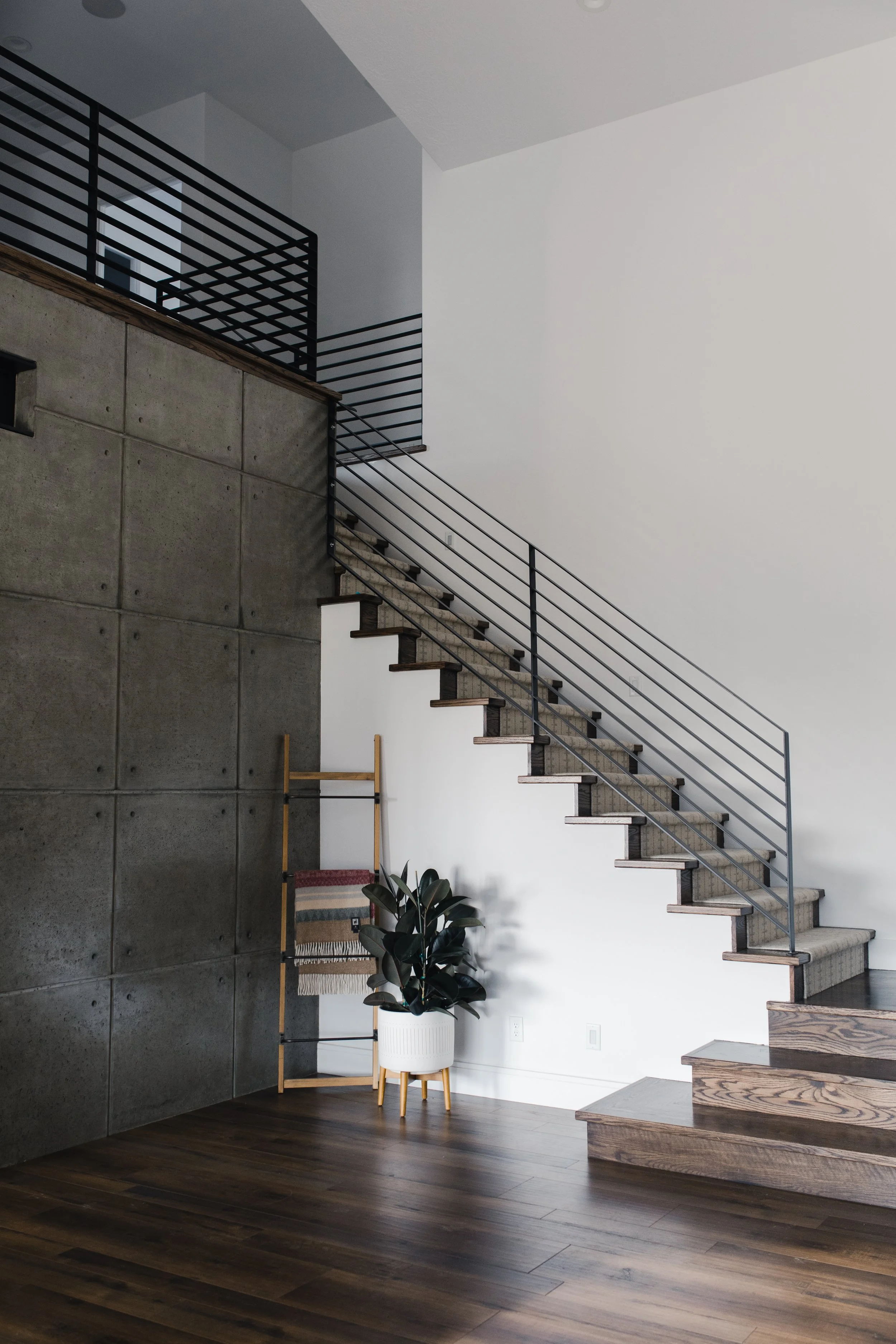 Modern staircase with dark wood steps and metal railing in a minimalist interior with white and concrete walls, a potted plant, and a decorative ladder with a blanket.