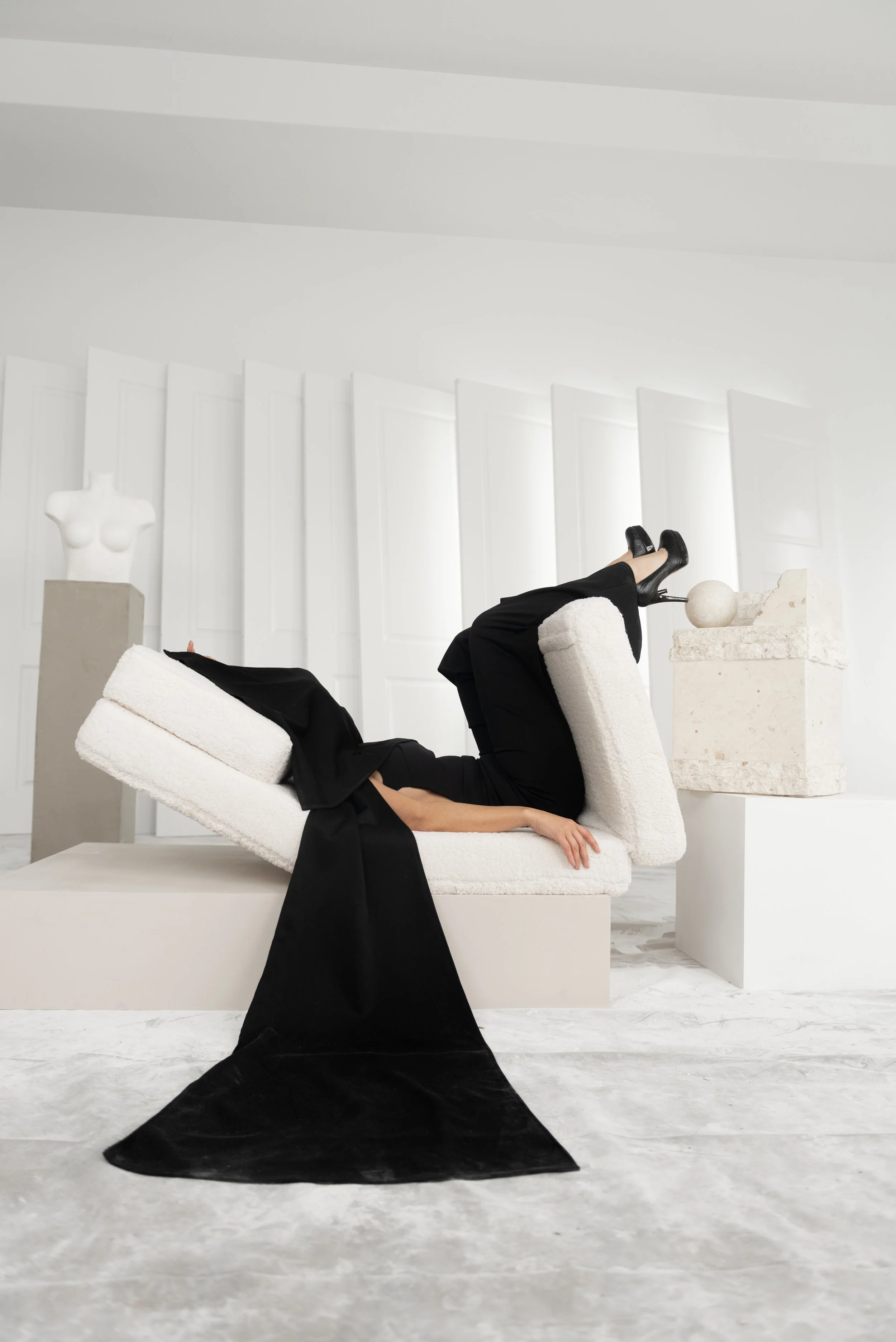 Person lying on a white, textured lounge chair in a minimalist, white art gallery or museum. They are dressed in black, with a black dress or coat and black high heels. The gallery features white sculptures and architectural elements, including a mal