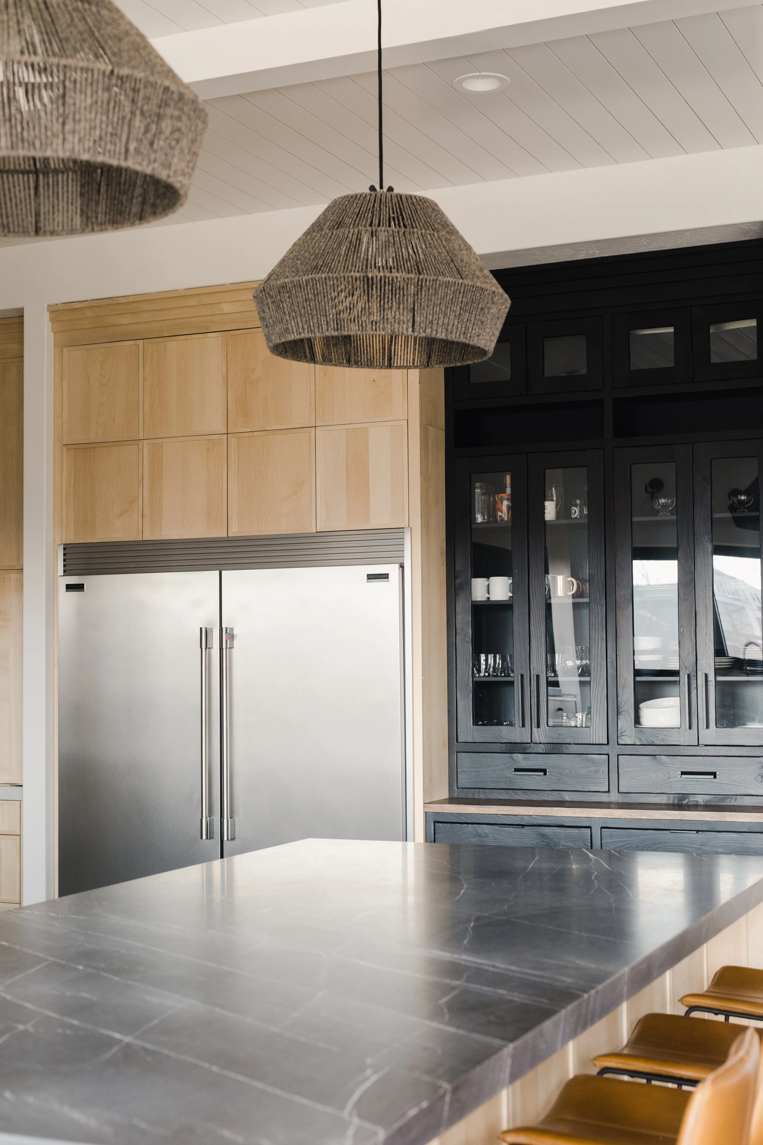 Modern kitchen with a gray marble countertop, stainless steel refrigerator, light wooden cabinets, and black glass-fronted cabinets displaying dishes, with woven pendant lights hanging from the ceiling.