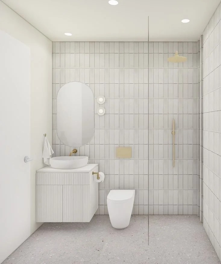 A modern minimalist bathroom with white tiled walls, a white toilet, a white vanity with a rounded vessel sink, oval mirror, wall-mounted light, and gold fixtures. The space features neutral tones and simple decor.