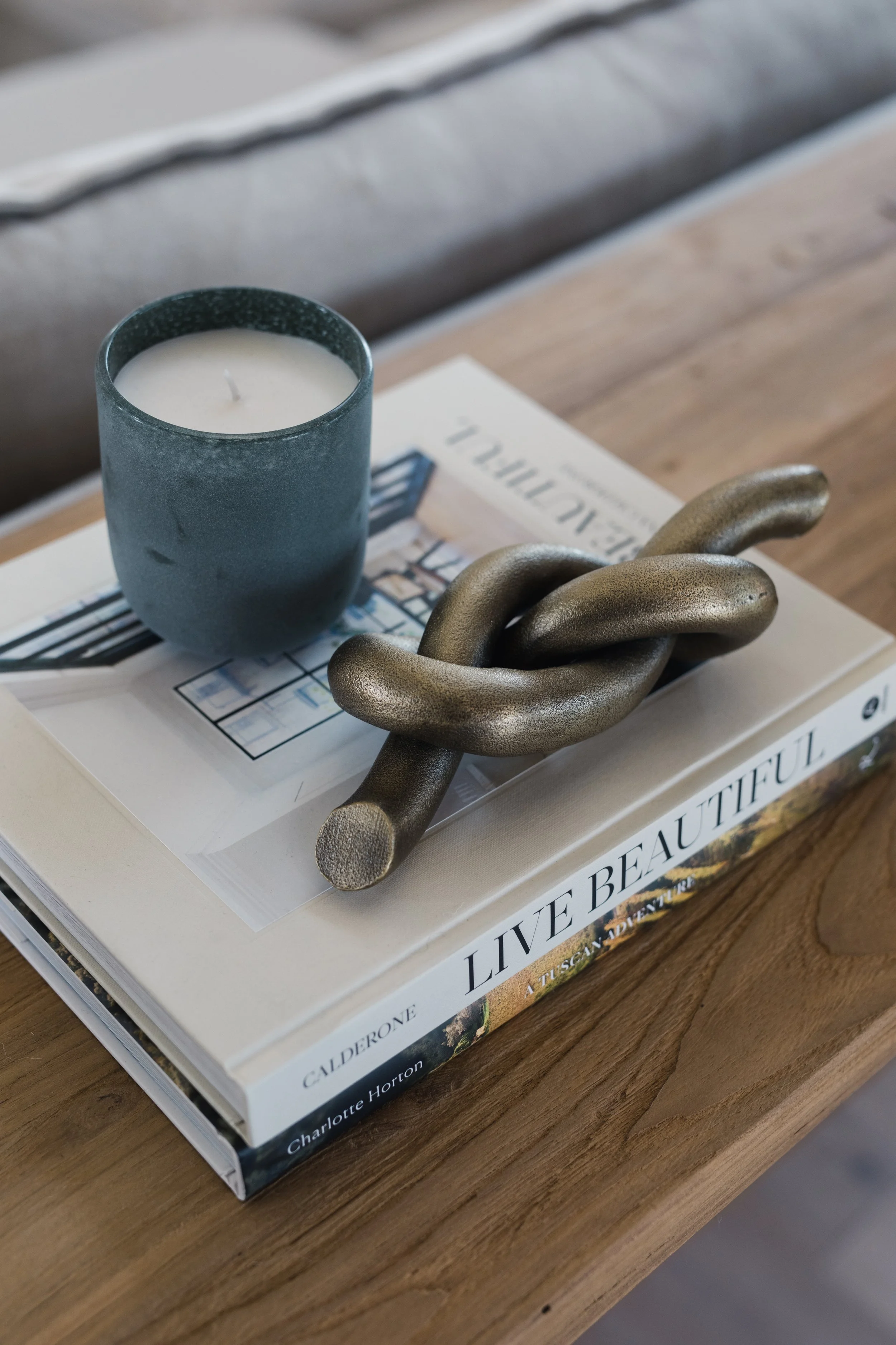 A bedside table with a blue candle, a decorative metallic chain sculpture, and a book titled "Live Beautiful" resting on a white magazine.