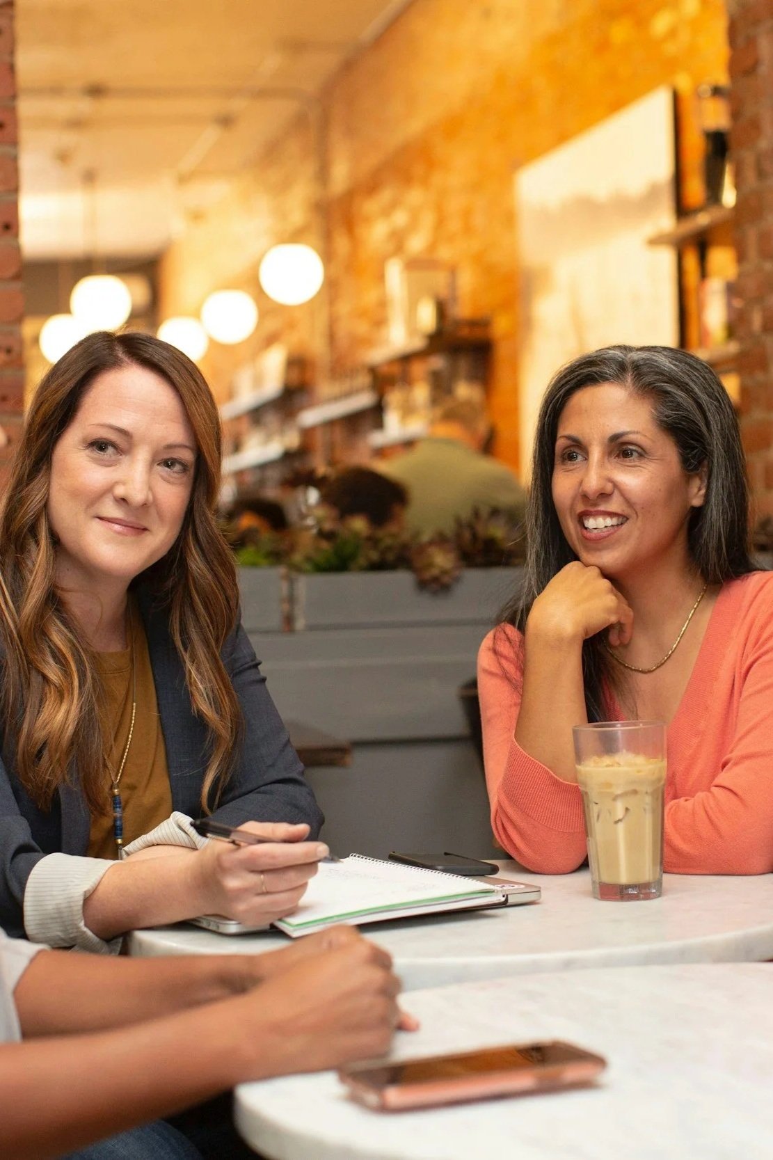 Two women, one with light skin and long brown hair, the other with brown skin and black hair streaked with gray. They are engaged in discussion and the light-skinned woman is taking notes. The background is blurred but appears to be a restaurant.