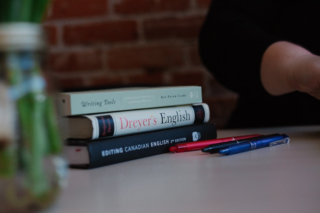 A stack of language and editing craft books is pictured against a red-brick background. The titles include: "Writing Tools," "Dreyer's English," and "Editing Canadian English." Pens in blue, black, and red lay alongside the books.