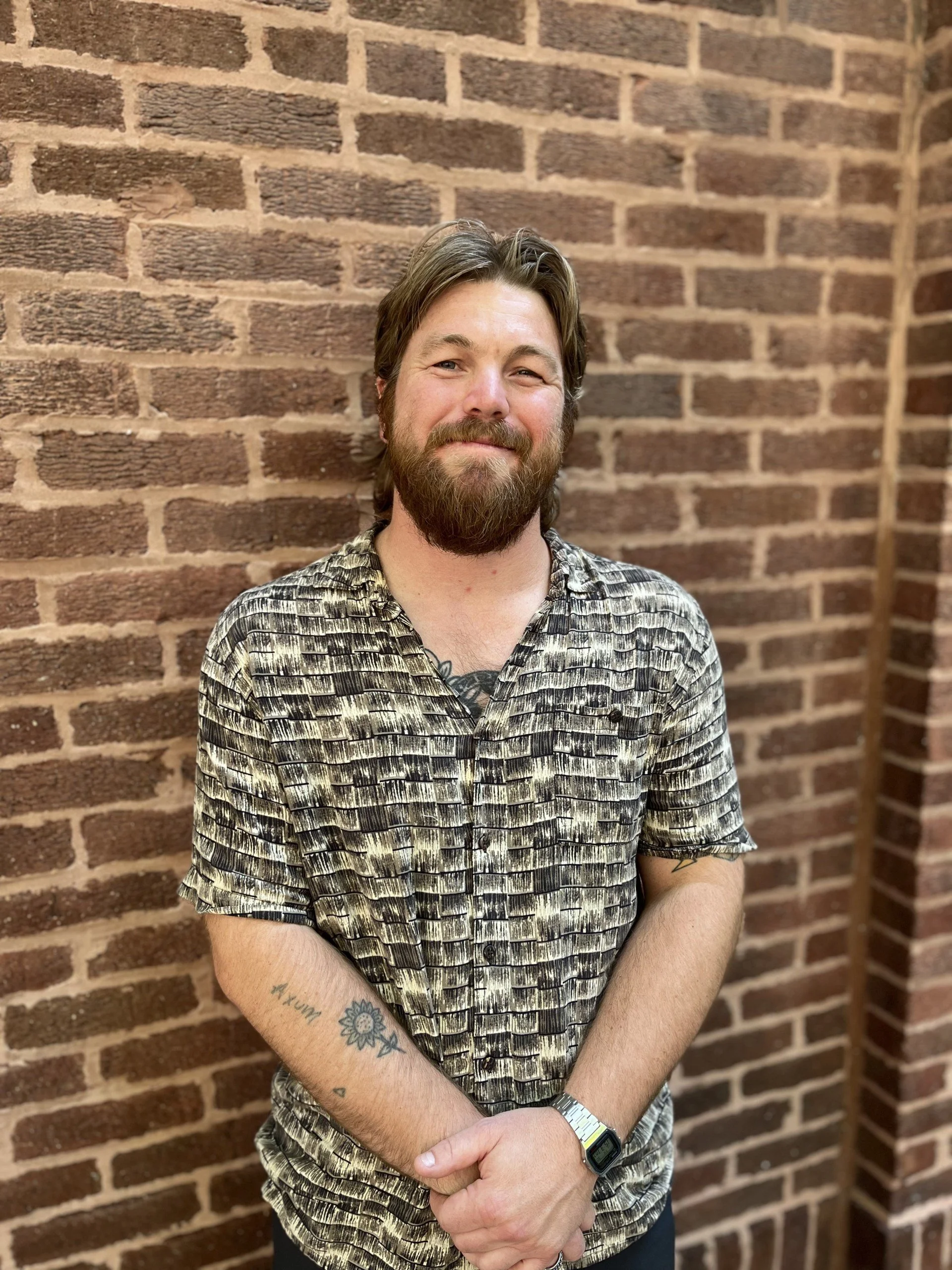 A man with a beard and tattoos standing in front of a brick wall, smiling slightly, wearing a patterned shirt and a digital watch.