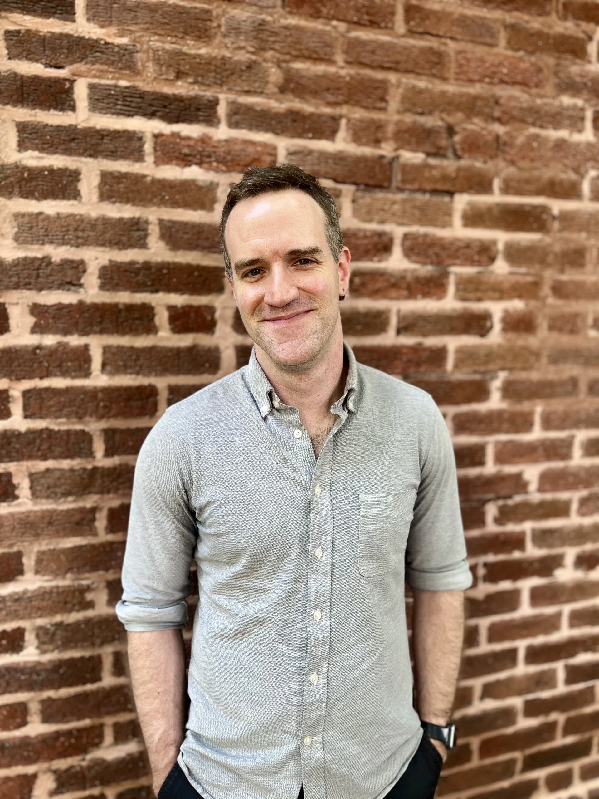 A man with dark hair and a light gray button-up shirt standing in front of a red brick wall, smiling slightly.